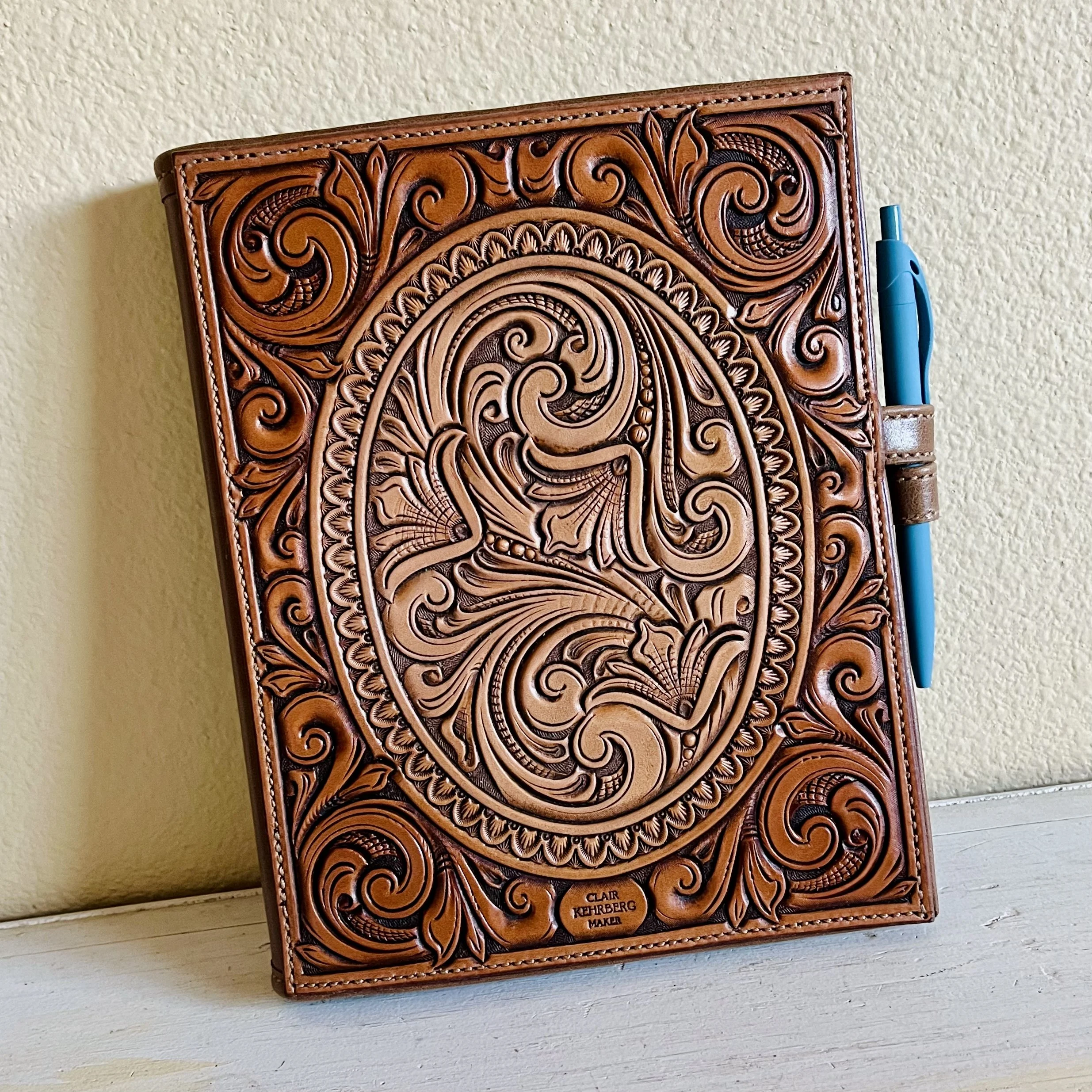 A leather-bound notebook with intricate carved designs on the cover, with a blue pen attached in a side loop.