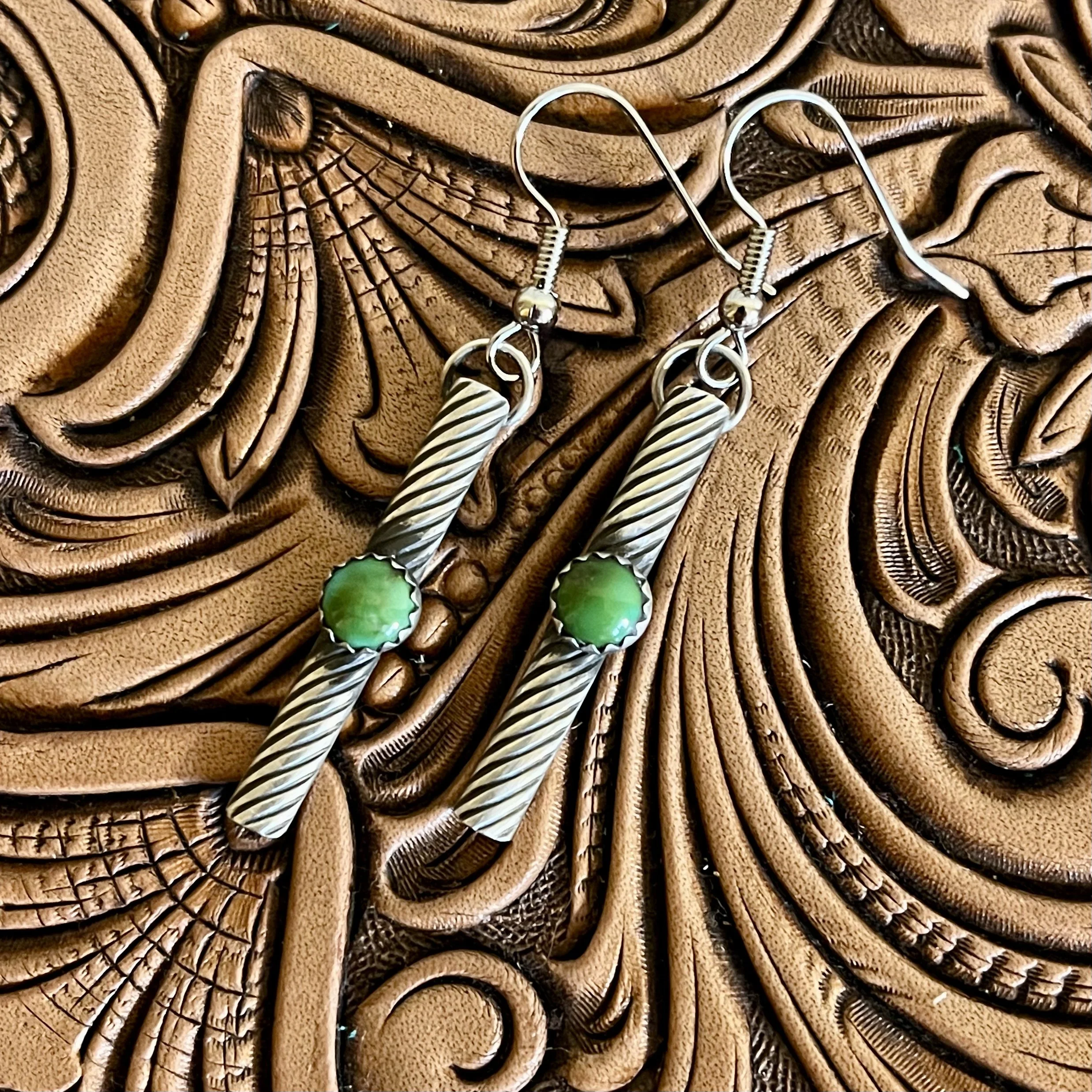 A pair of silver earrings with green stones resting on an intricately carved leather surface.