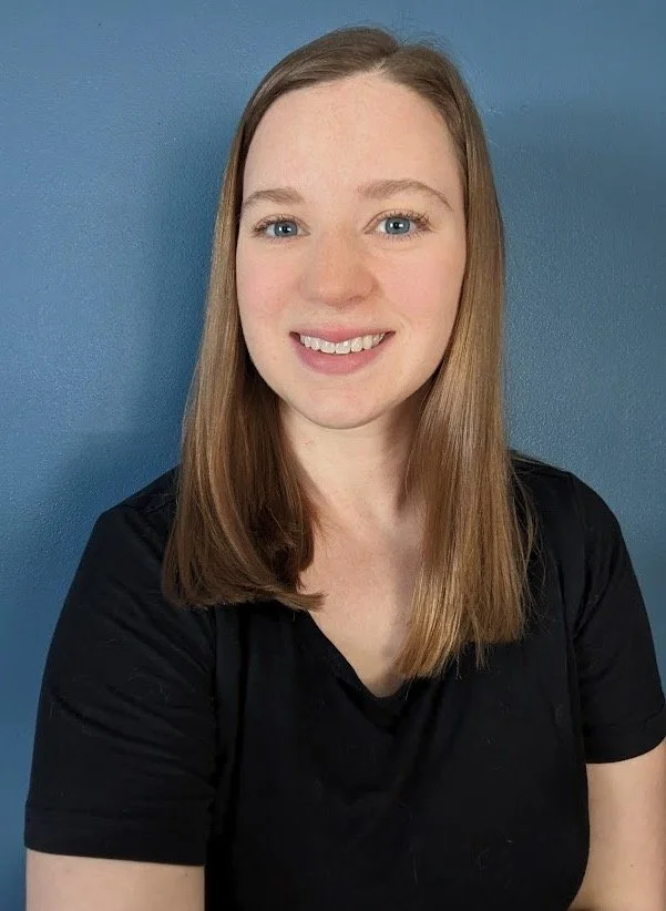 A smiling woman with long light brown hair, blue eyes, wearing a black shirt, standing against a blue wall.