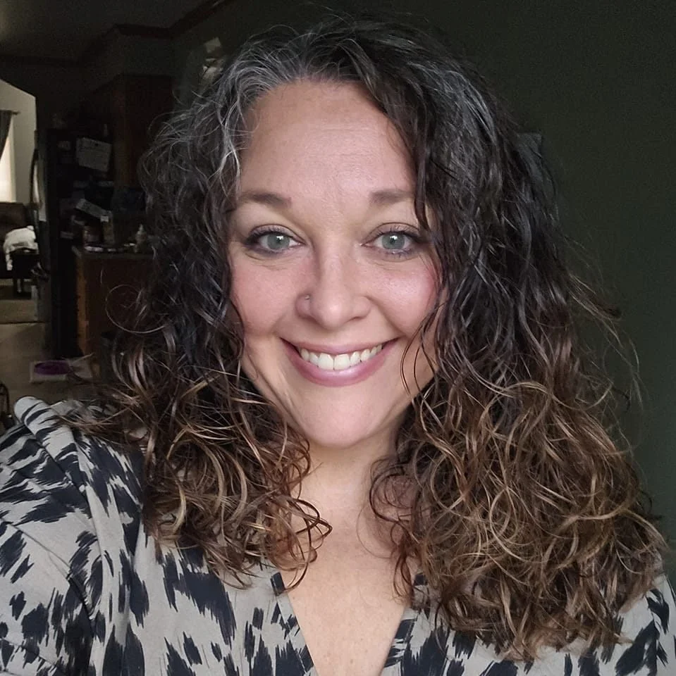 A woman with curly brown hair and blue eyes smiling at the camera in a home setting.