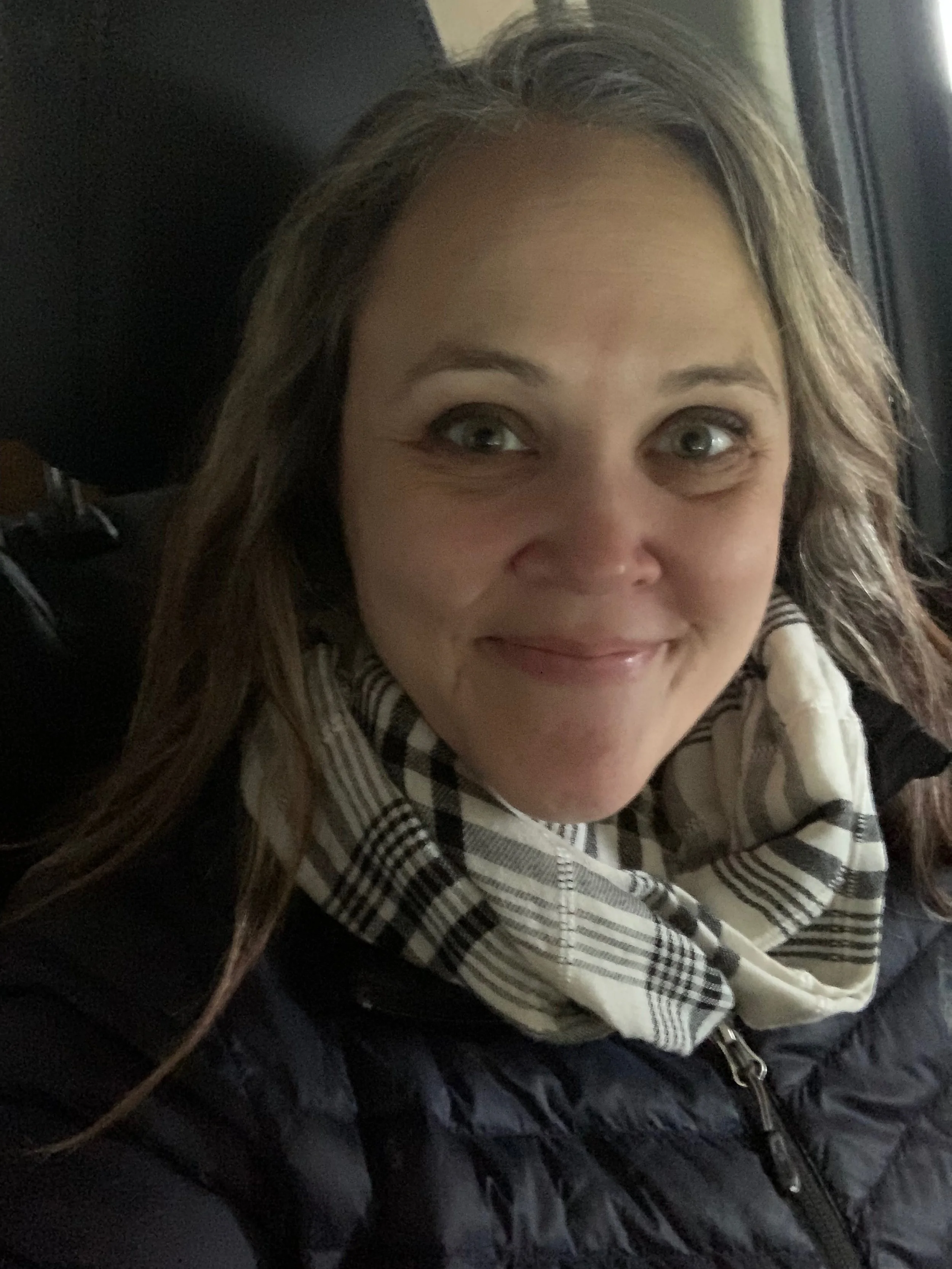 A woman with long brown hair, wearing a black quilted jacket and a black and white checkered scarf, smiling and sitting near a window.