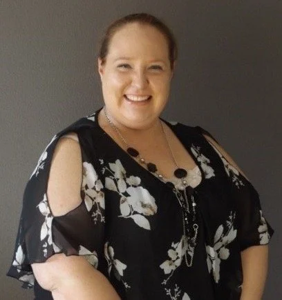 A smiling woman with light skin and brown hair tied back, wearing a black floral dress with cut-out shoulders, standing against a gray background.