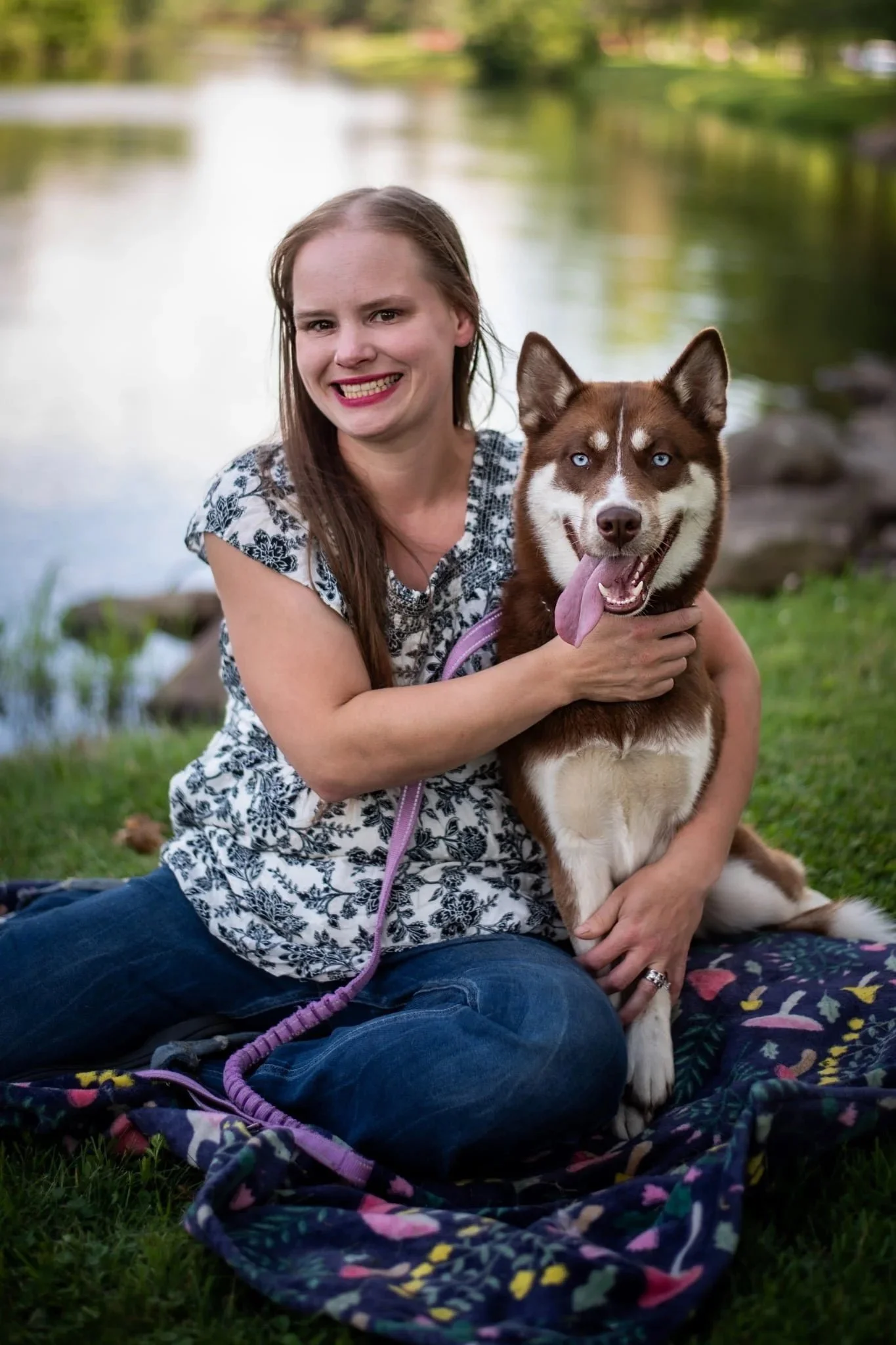 A woman sitting on the grass by a lake, holding a Siberian Husky dog with blue eyes and sticking out tongue.