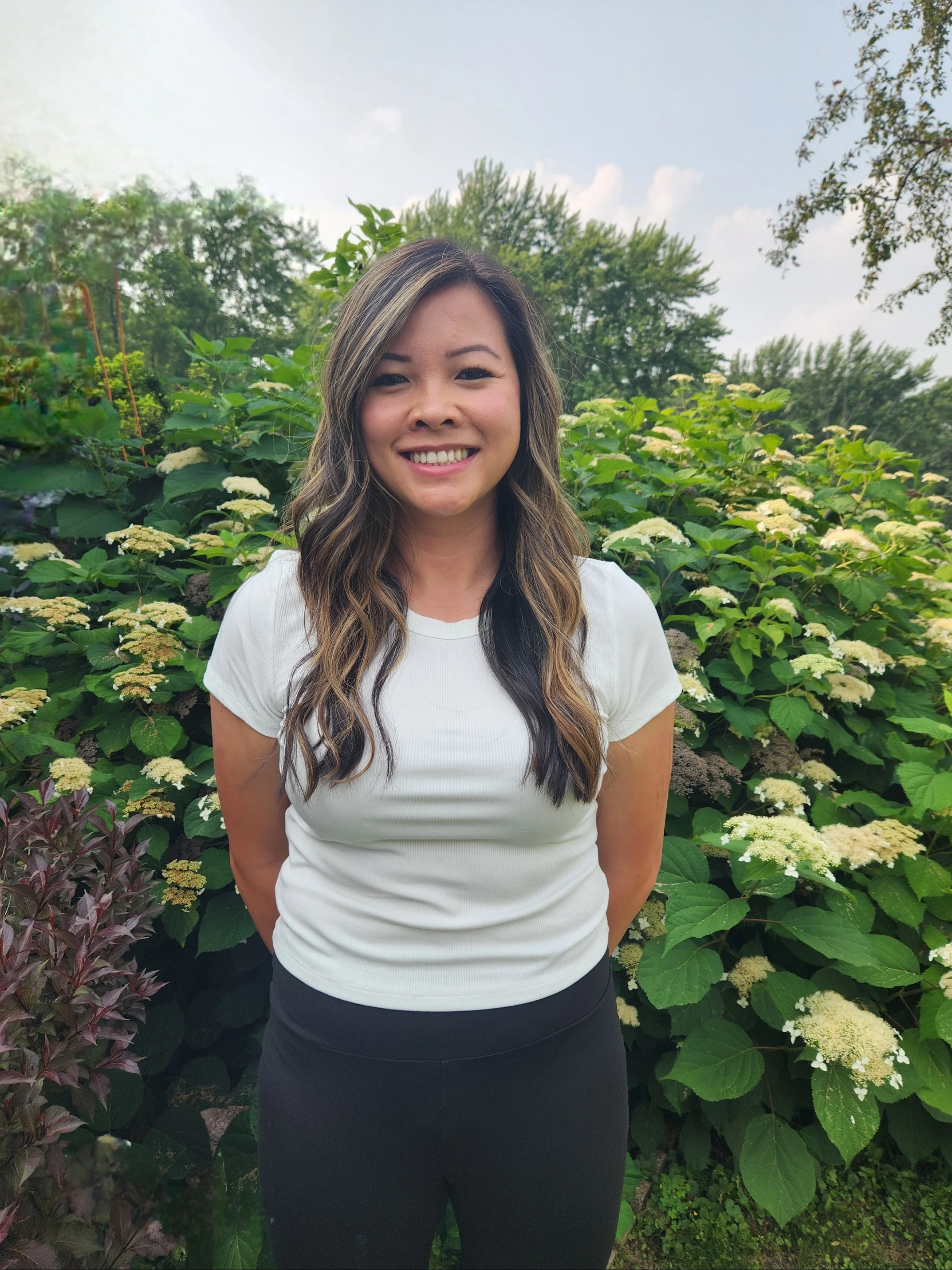 Smiling woman in white shirt standing outdoors in front of lush green bushes and trees.