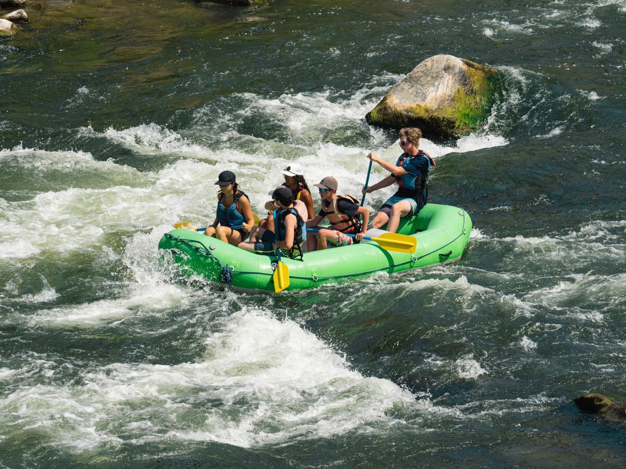 A green raft with passengers navigates mild whitewater.
