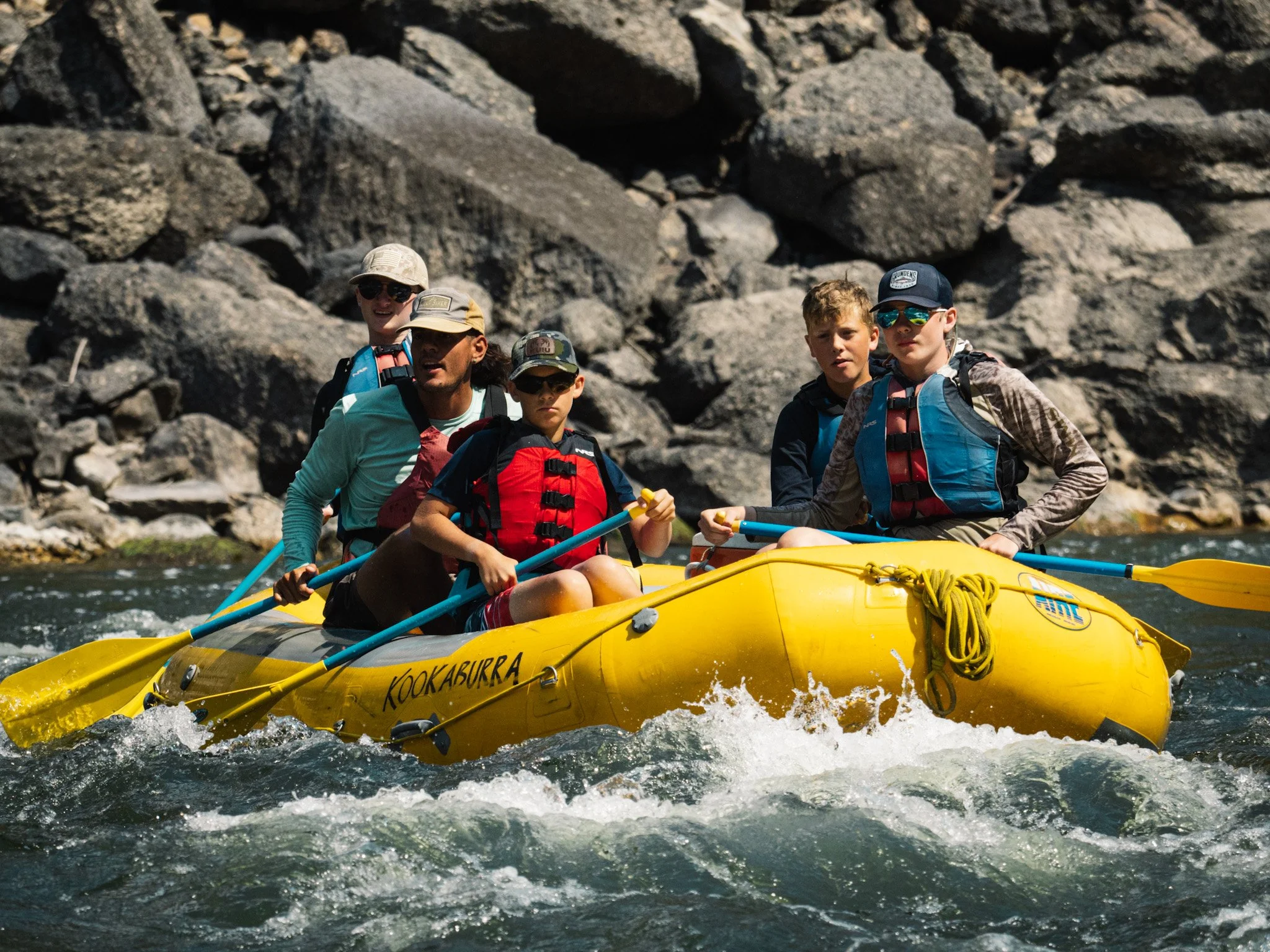 Passengers in a yellow raft focus their attention downstream as they float through waves in a river with large rocks in the background.