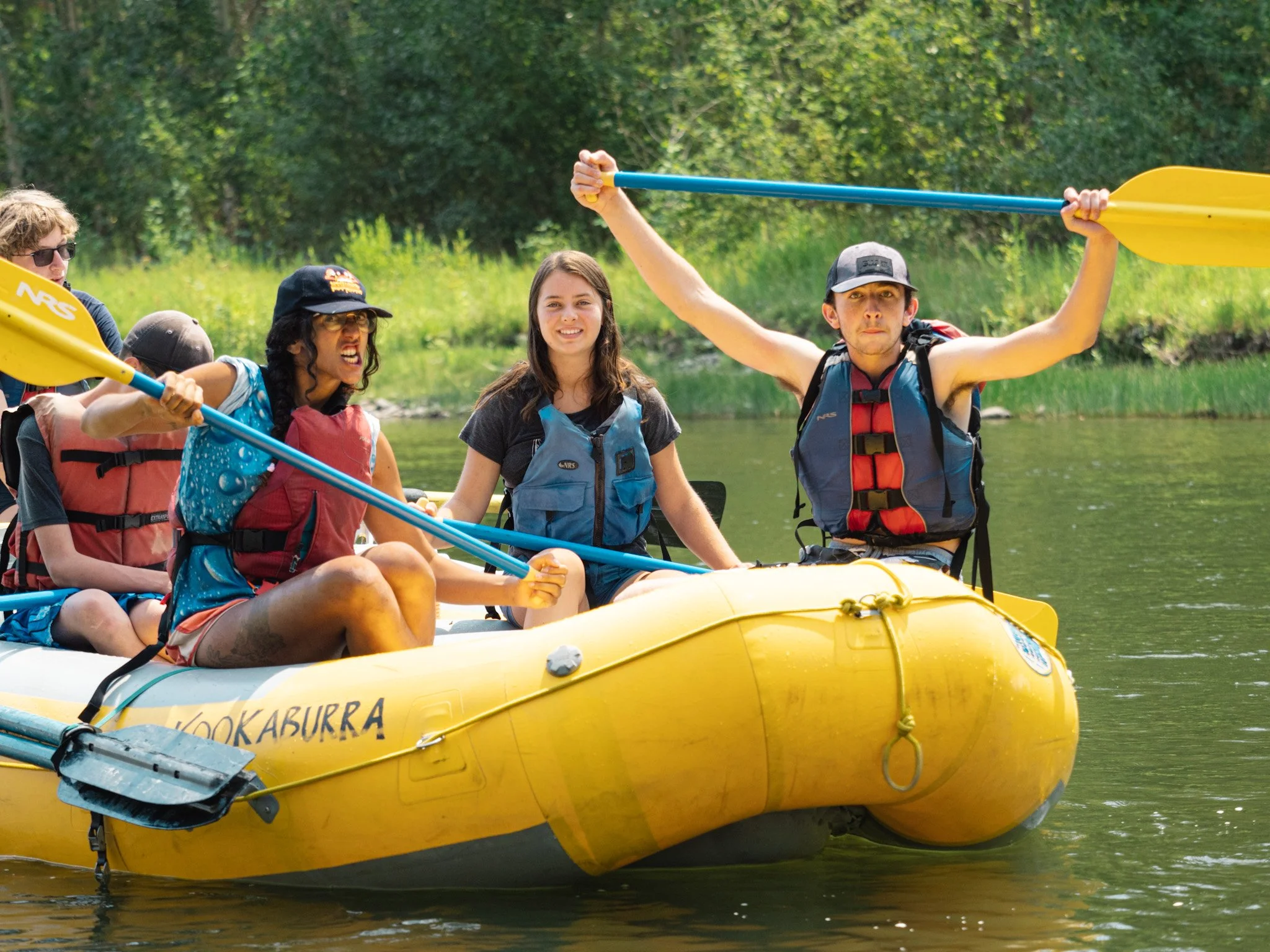 A passenger in the front of a raft holds their paddle over their head.