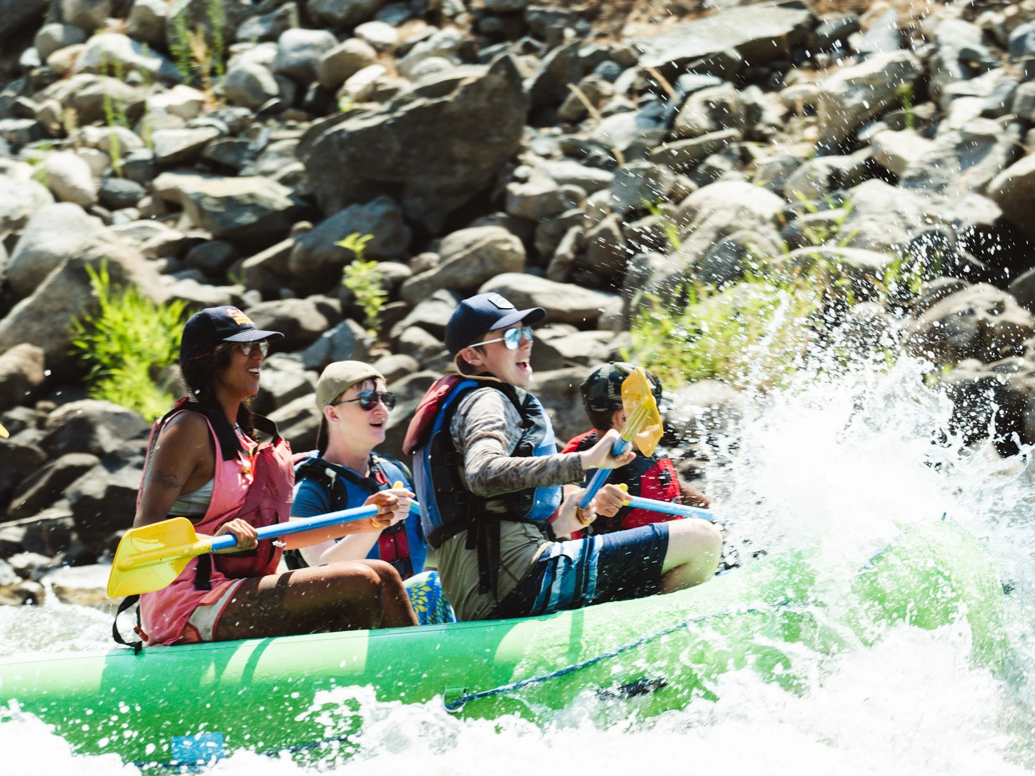 A green raft punches through a wave in a river sending water splashing over the bow of the boat and onto the passengers.