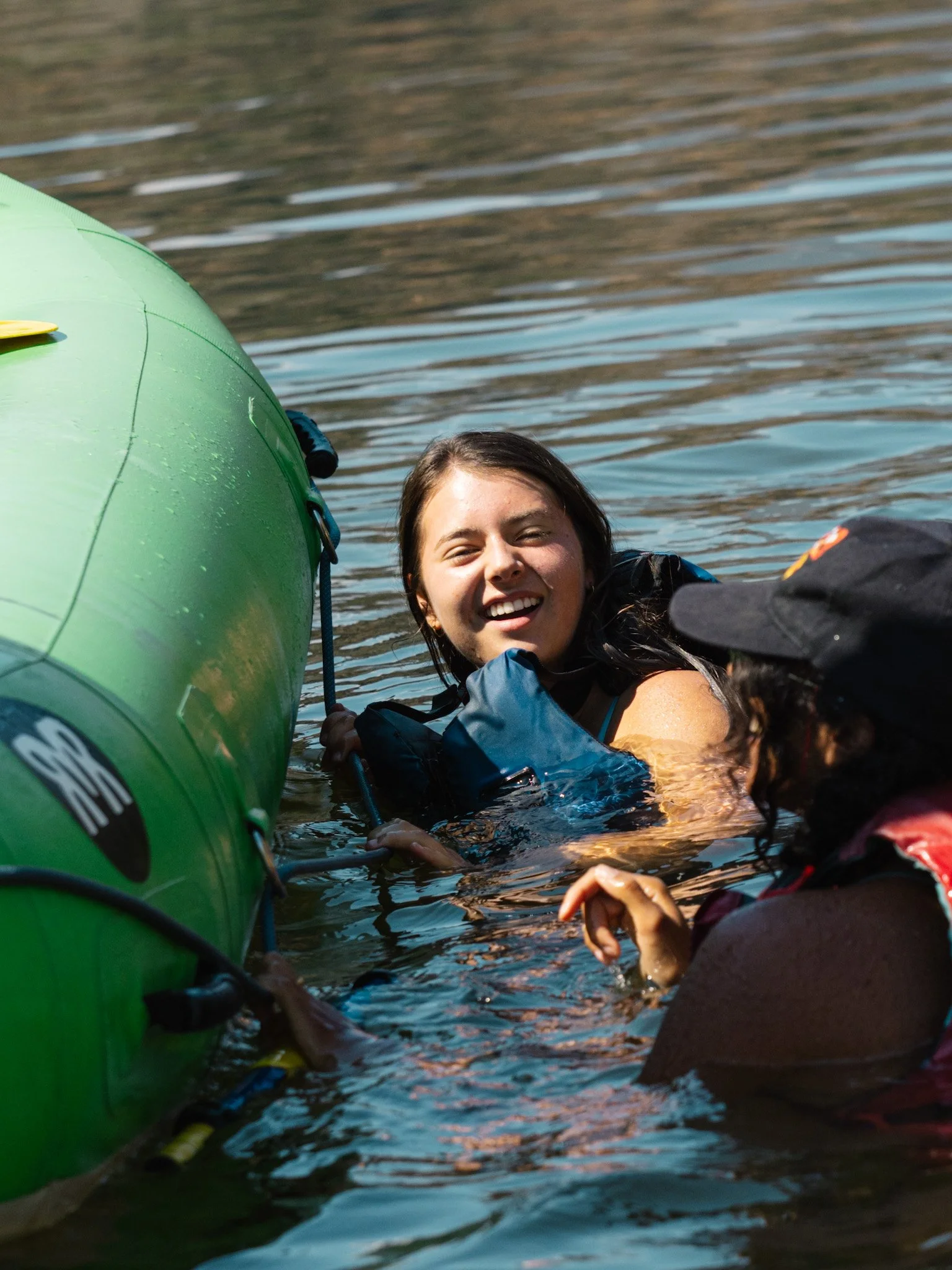 Two students swim alongside of a green raft in calm water.