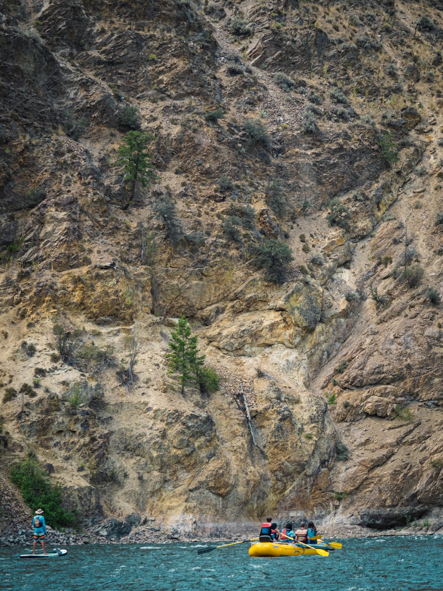 A standup paddle boarder and a raft approach a tall bluff while floating down a river.