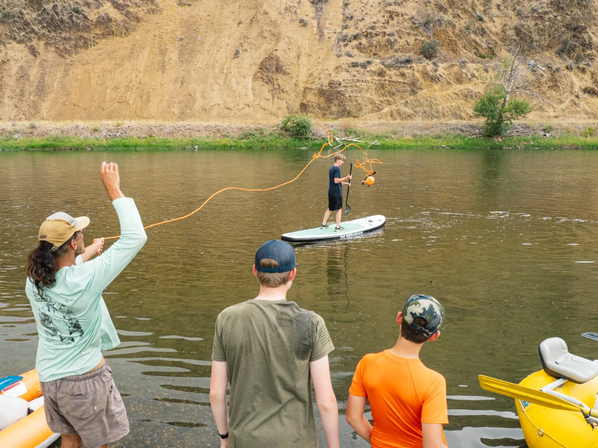 A river guide throws a rescue line to a camper on a paddle board during a river rescue training session while several other campers watch.