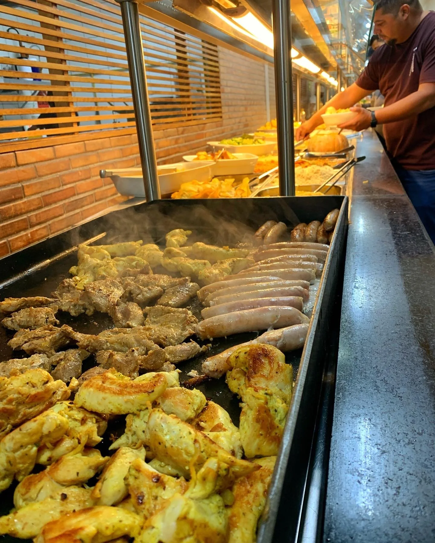 Churrasqueira com carnes variadas e linguiças em um restaurante, com um homem servindo-se ao fundo.