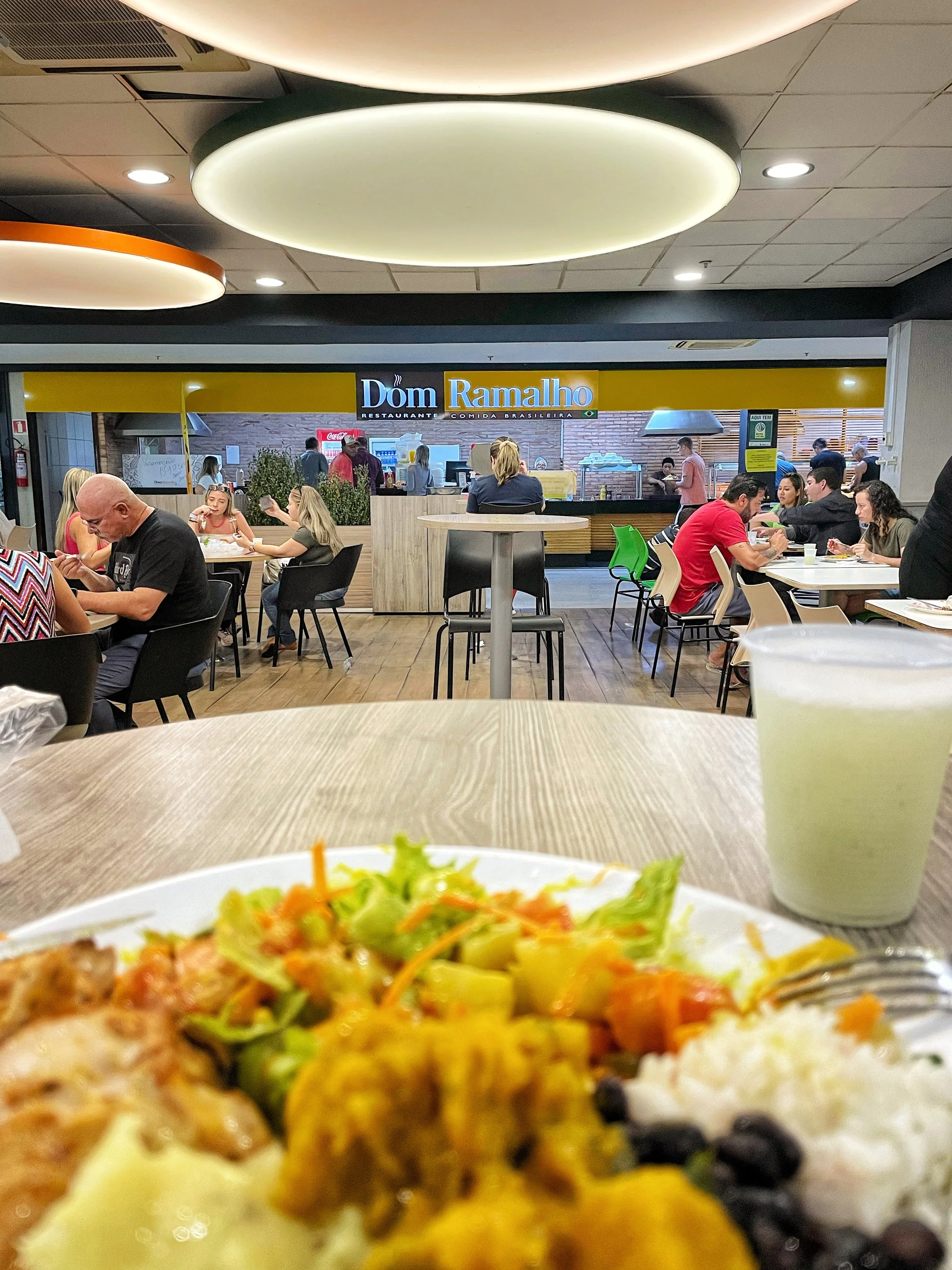 Interior de restaurante com várias pessoas em mesas, um prato de comida brasileira em primeiro plano e um copo de bebida ao lado. Logo "Dom Ramalho" visível ao fundo.