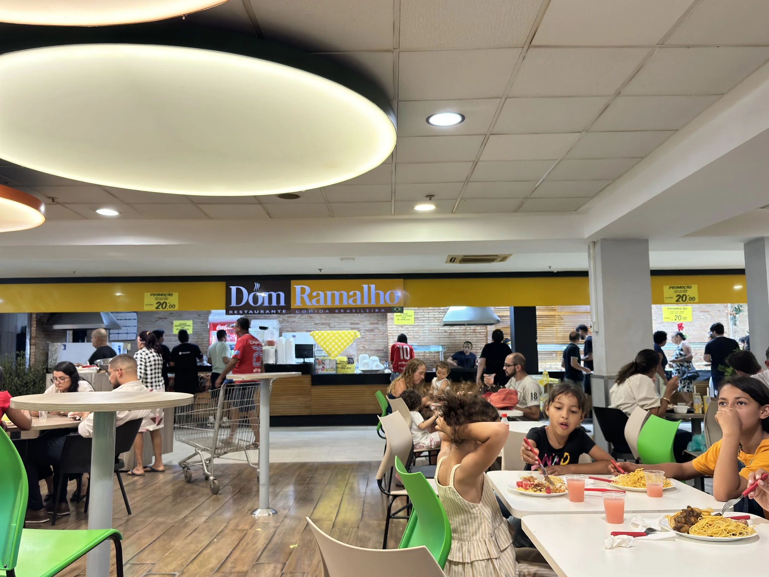 Interior de restaurante com pessoas consumindo refeições, incluindo crianças na mesa com pratos de massa, e uma área de atendimento com sinalização de promoções.
