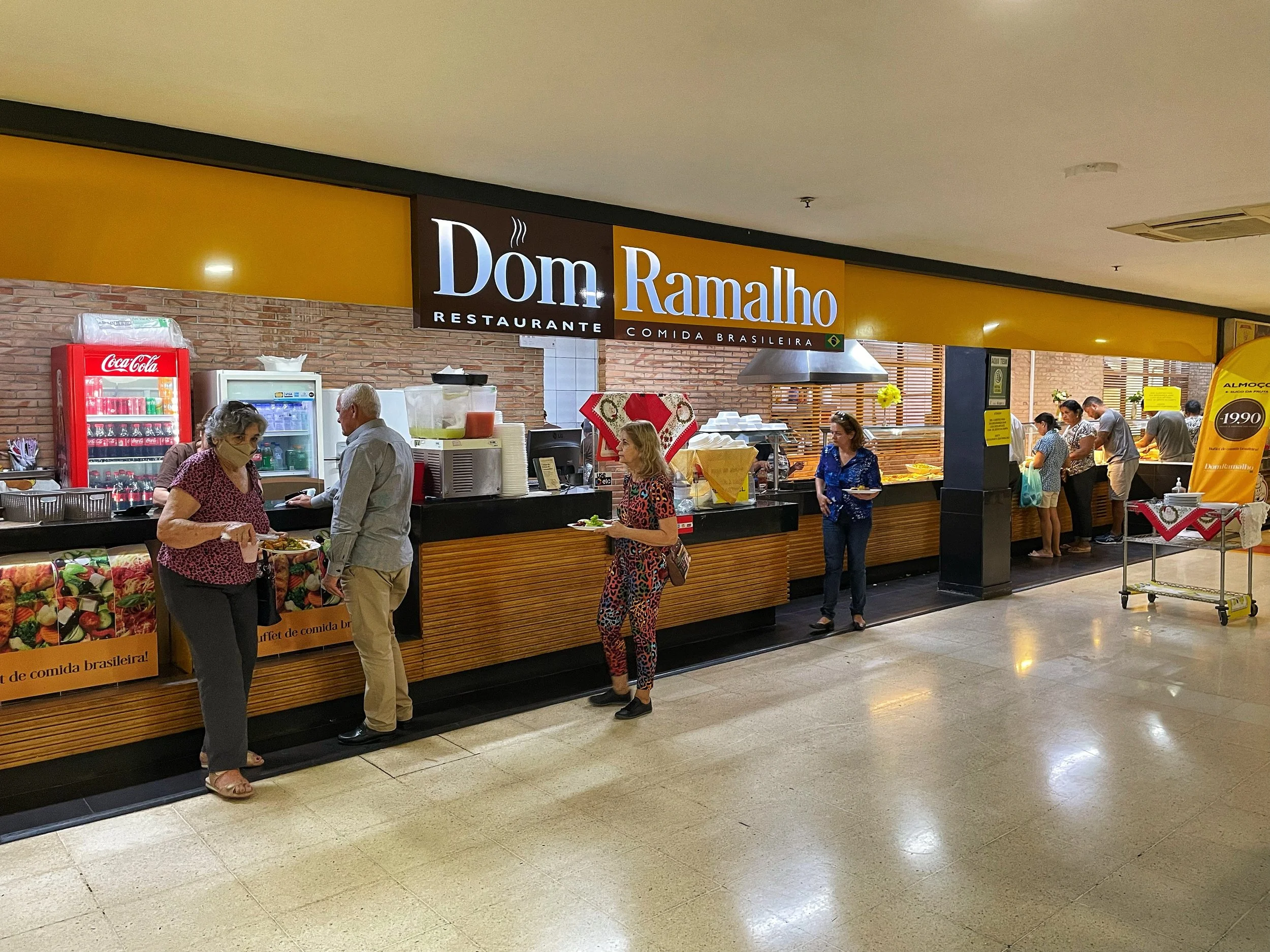 Clientes servindo-se em um restaurante de buffet chamado 'Dom Ramalho', especializado em comida brasileira, com uma fachada amarela e bebidas expostas ao fundo.