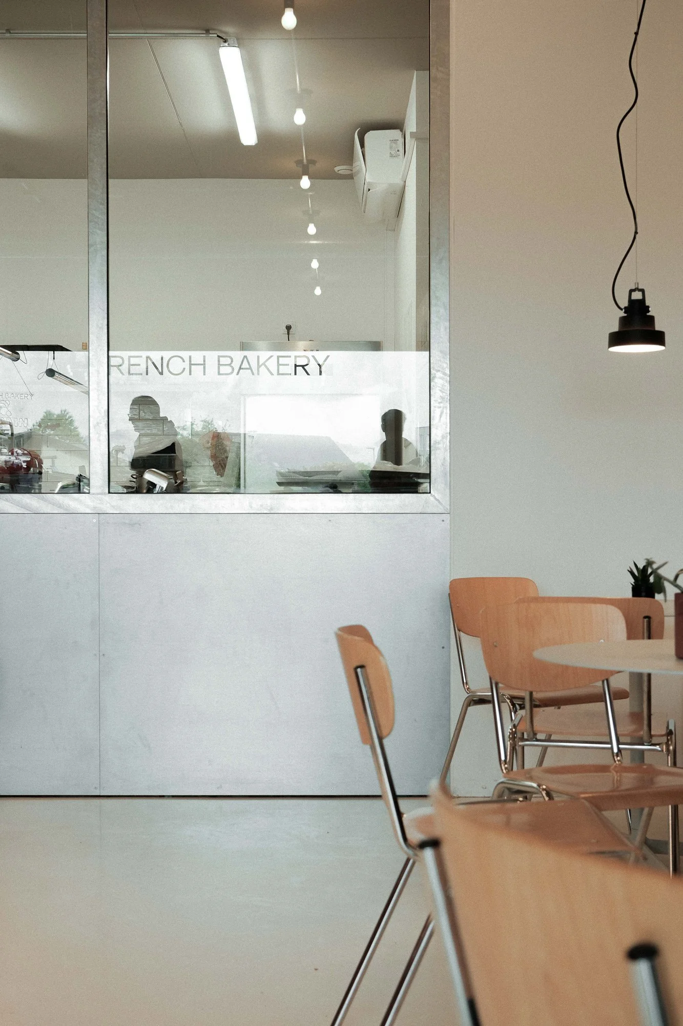 Einblick in eine Bäckerei mit Glasfenster, in dem zwei Silhouetten von Personen zu sehen sind, und einem Raum mit Holztischen und Stühlen.