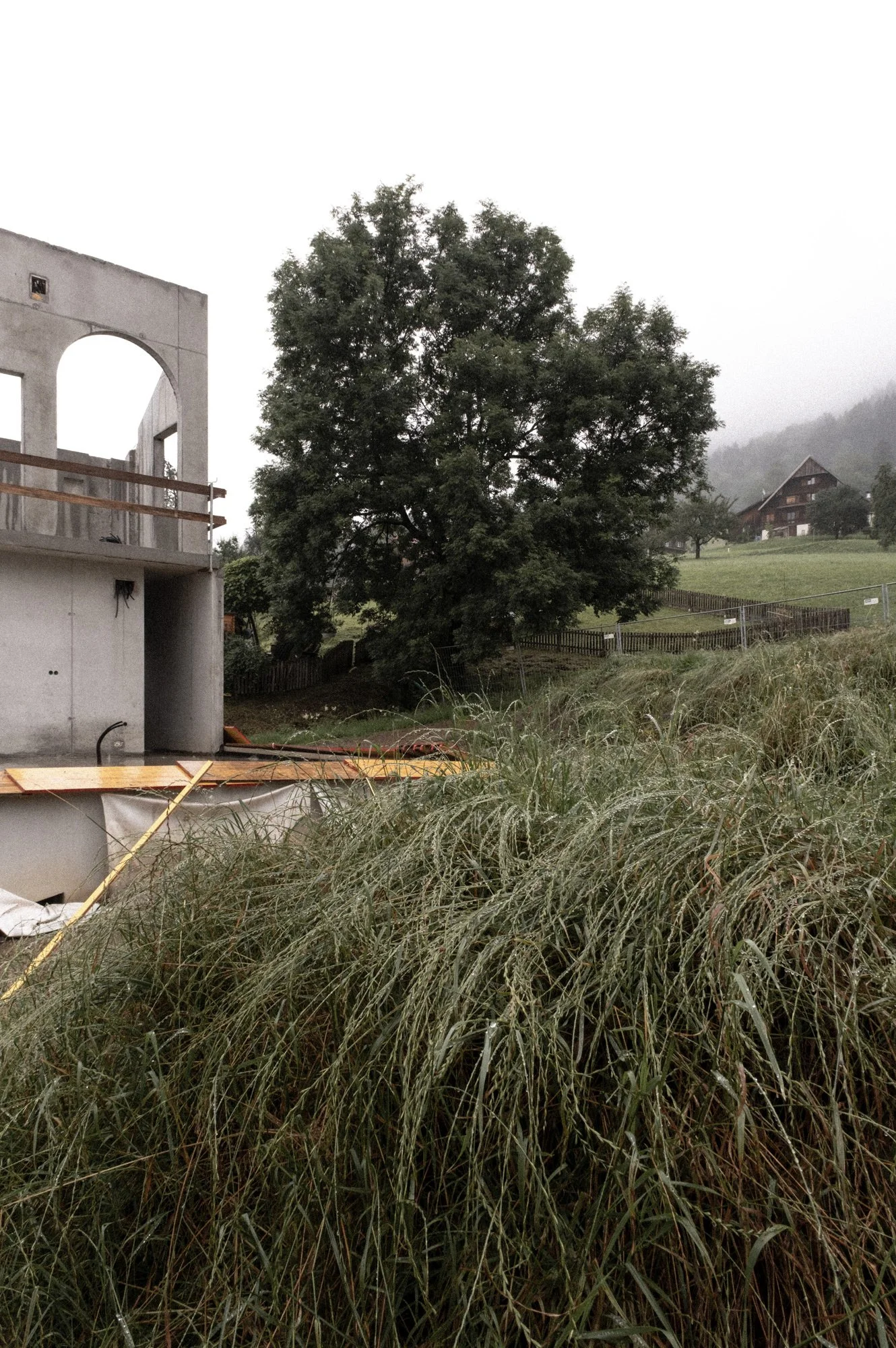 Ein Baustellenbereich mit unvollendeter Gartenmauer, hohen Gräsern im Vordergrund, einem großen Baum, einer dichten Nebeldecke und einem Haus im Hintergrund auf einer hügeligen Landschaft.