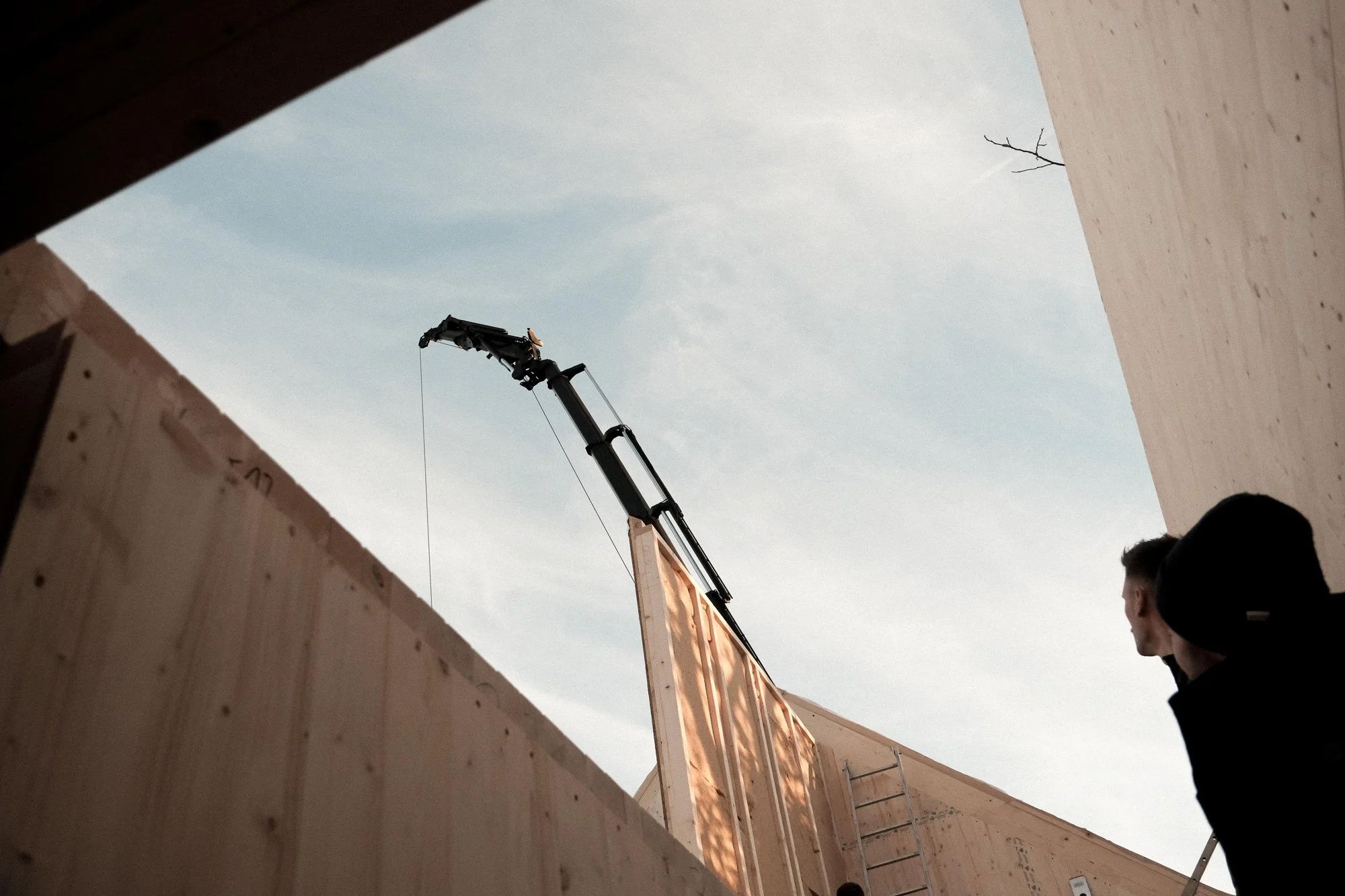 Blick durch eine bauliche Öffnung auf eine Baukran, der an einem Holzrahmen im Gebäude arbeitet, mit zwei Männern im Vordergrund, einer mit schwarzer Kapuzenjacke, blickt nach oben bei Sonnenlicht.