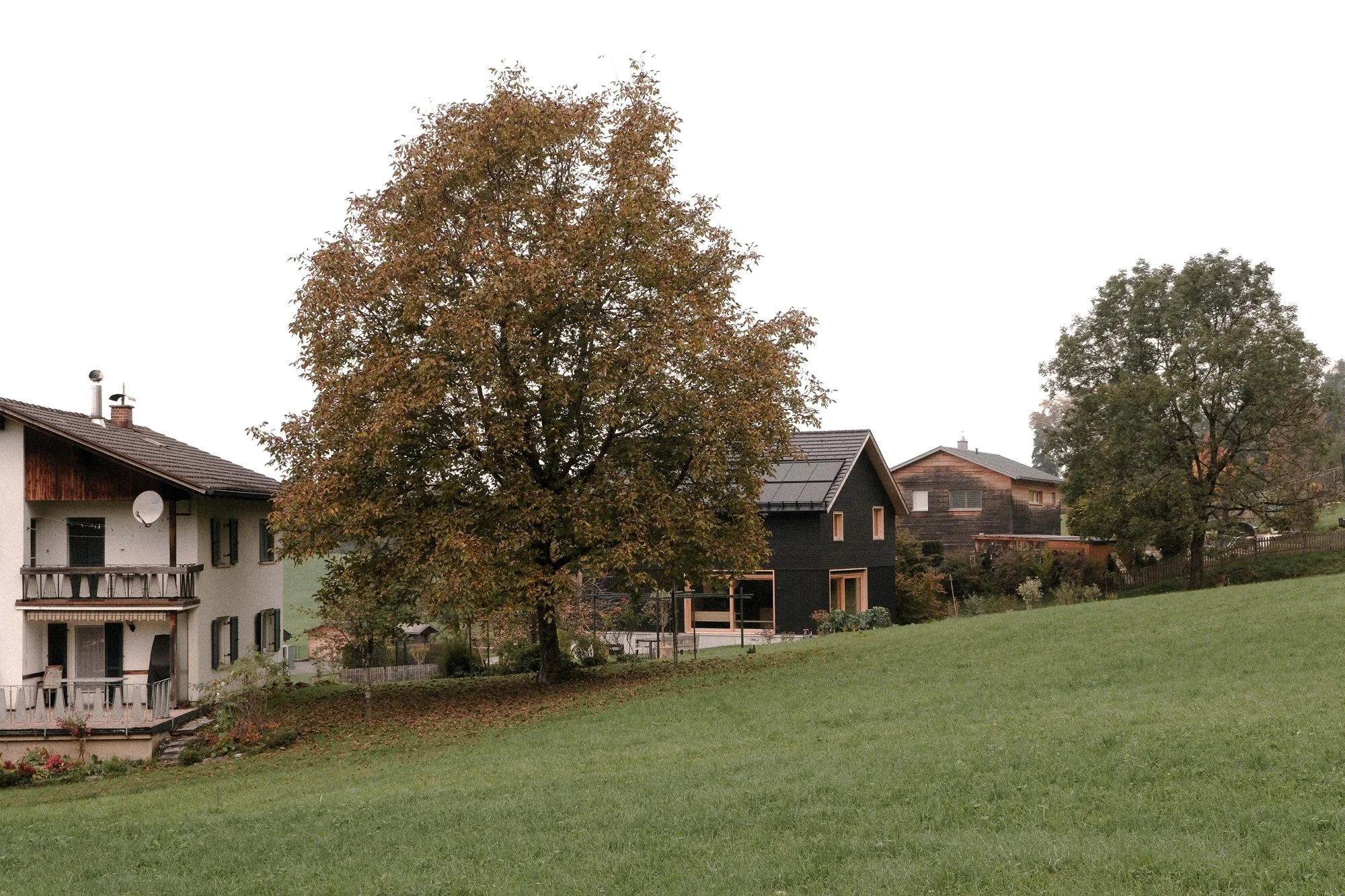 Ein ländliches Haus mit Balkon und Satellitenschüssel, umgeben von Bäumen und einer grünen Wiese, bei bewölktem Himmel.