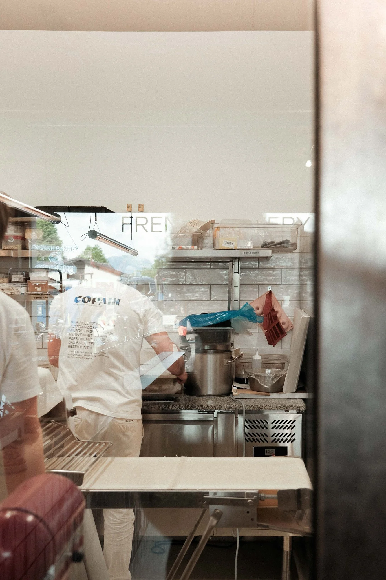 Blick durch eine Glasscheibe in eine professionelle Küche, in der Kocharbeiten ausgeführt werden. Reflexionen und Silhouetten der Köche sind sichtbar.