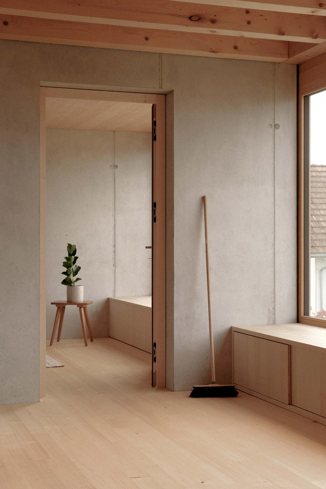 Innenraum eines minimalistischen Raums mit Holzfußboden, einer kleinen Holzbank mit Pflanzen, einem Fenster und einem Besen.