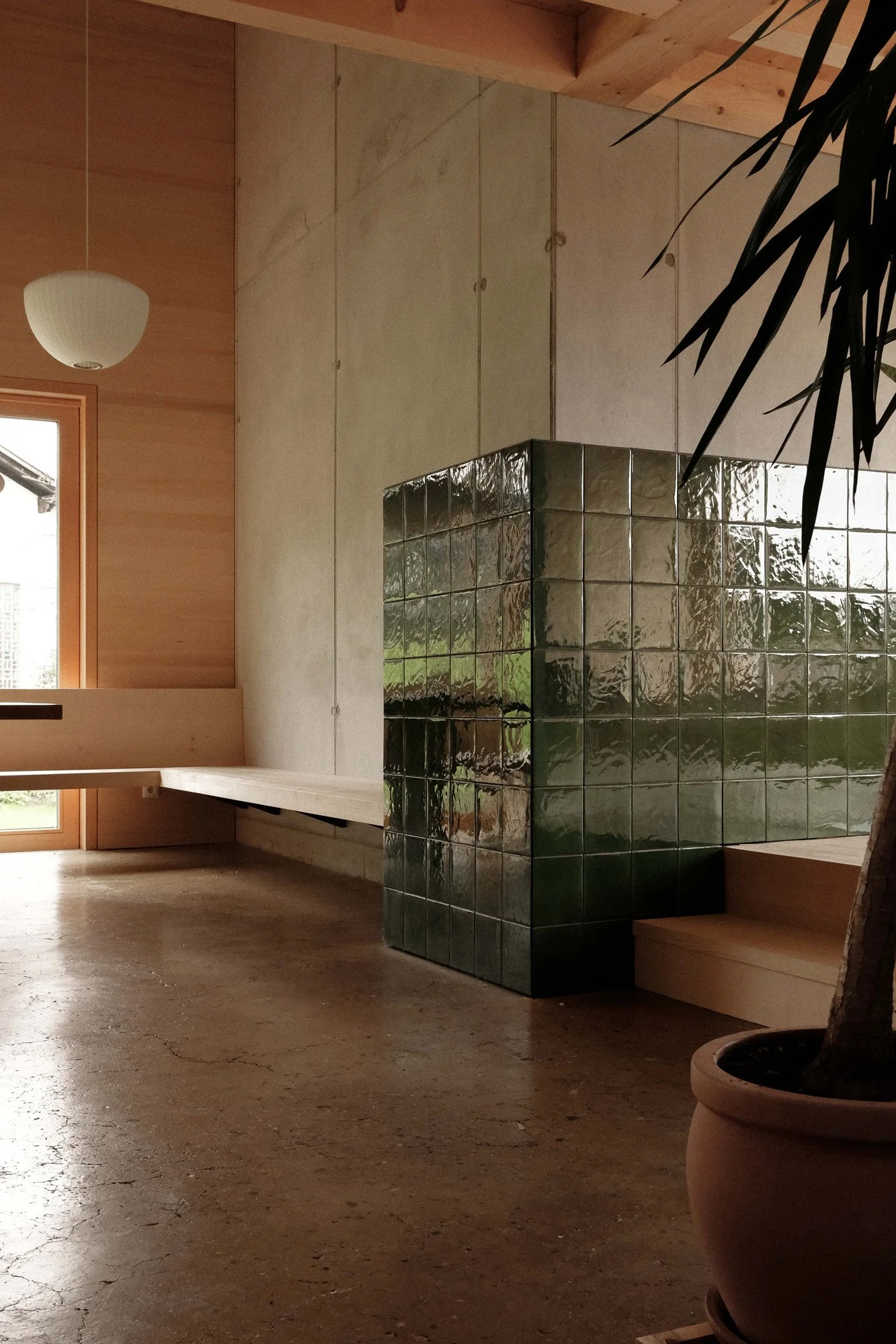 Innenraum mit natürlichen Holzmöbeln und grüner keramischer Ofen, große Fenster, Pflanzen, und Holzboden.