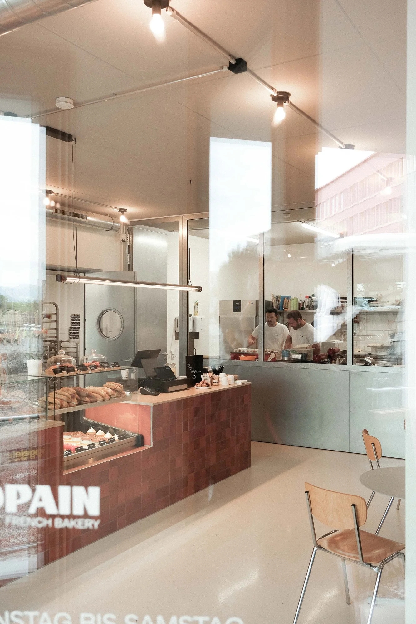 Blick in ein modernes französisches Bäckerei mit zwei bekannten Bäckern hinter der Theke, im Vordergrund eine Vitrine mit Gebäck, rechts zwei leere Tische mit Stühlen.