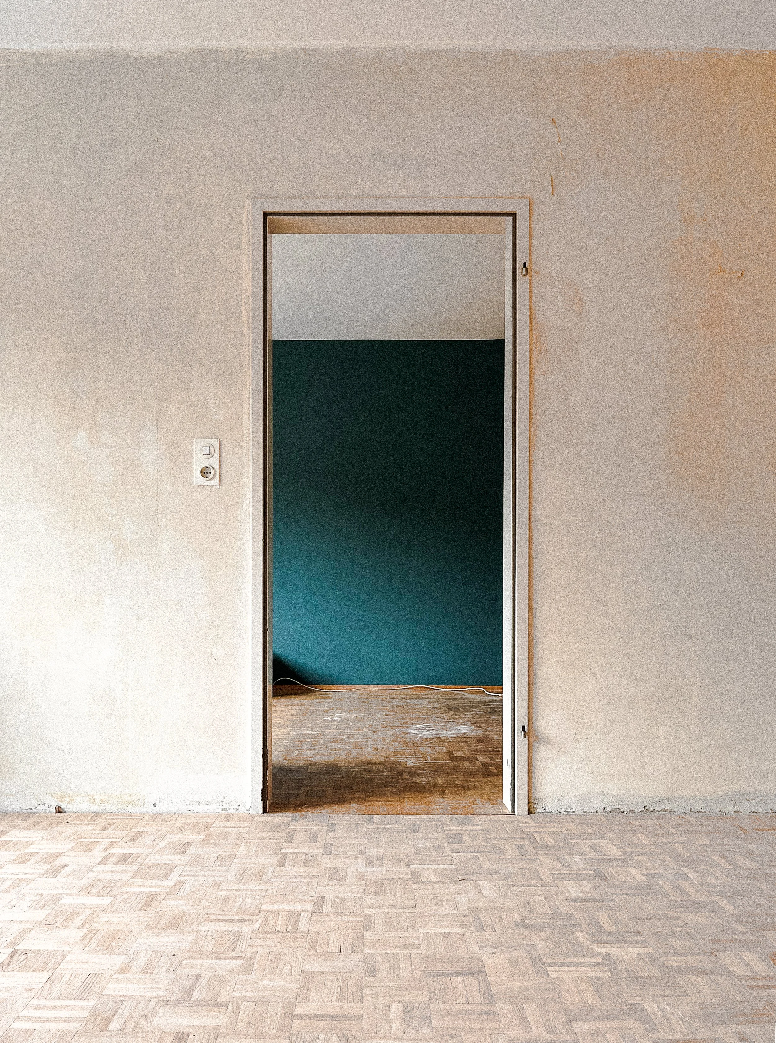 Leere Zimmer mit offenem Türrahmen, weißer Wand und Holzboden, im Hintergrund eine dunkelgrüne Wand in einem anderen Raum.