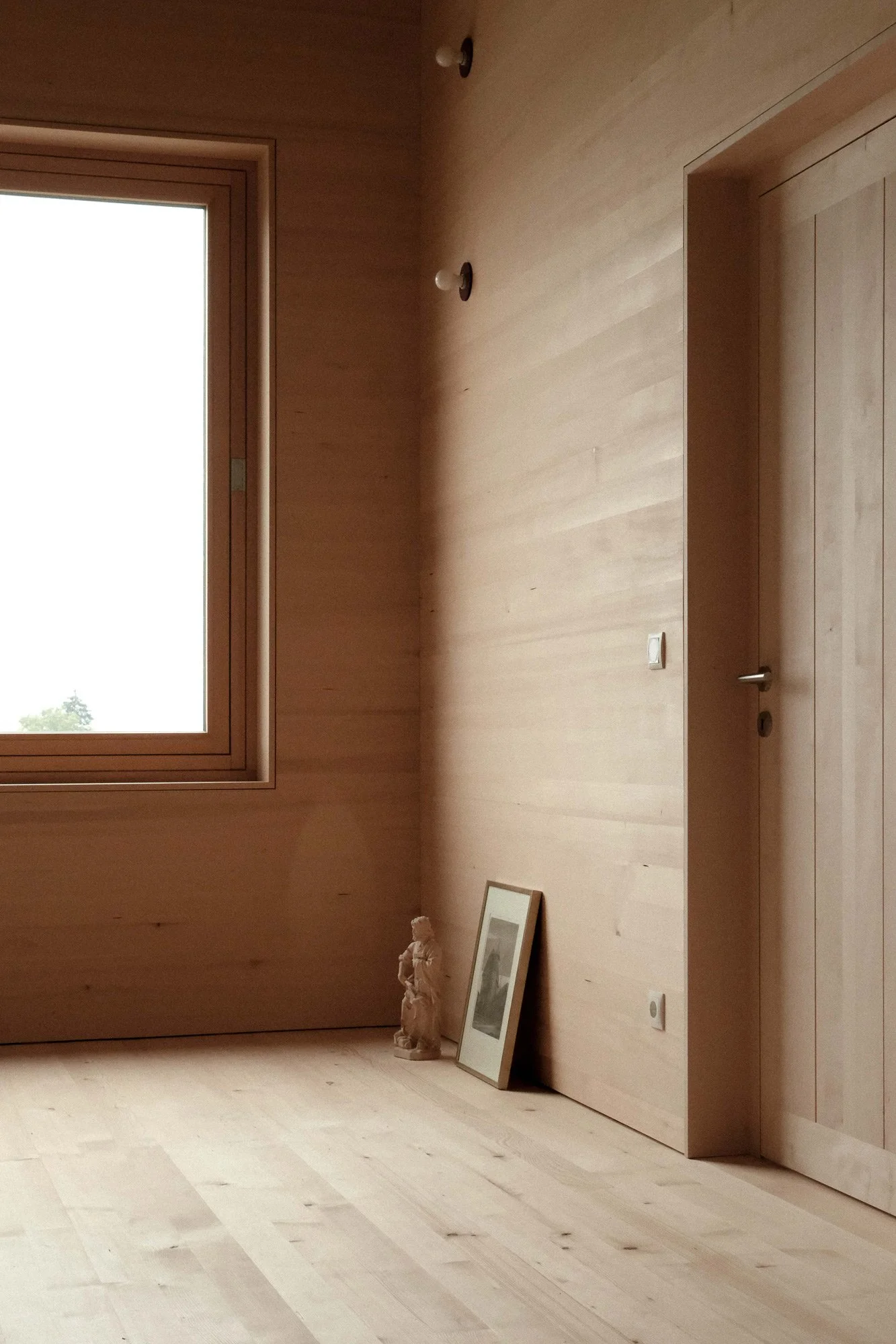 Ein Raum mit Holzwänden, einem Fenster auf der linken Seite, zwei Deckenleuchten, ein liegendes Bild und eine Skulptur im Ecken auf dem Boden.