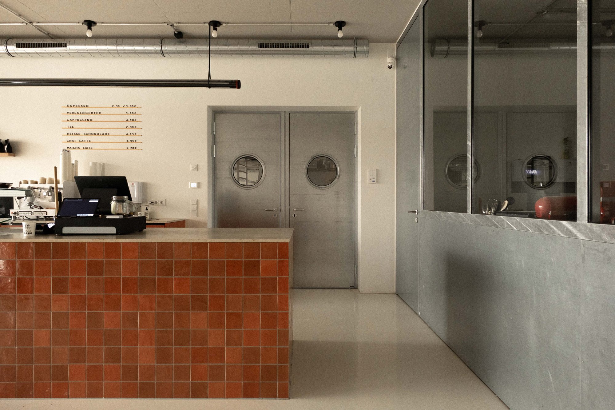 Innenansicht eines Cafés oder Coffeeshops mit einer Theke aus roten Fliesen, einem Kassensystem, einer Tür mit kleinen runden Fenstern und einer Fensterwand aus Metall und Glas.
