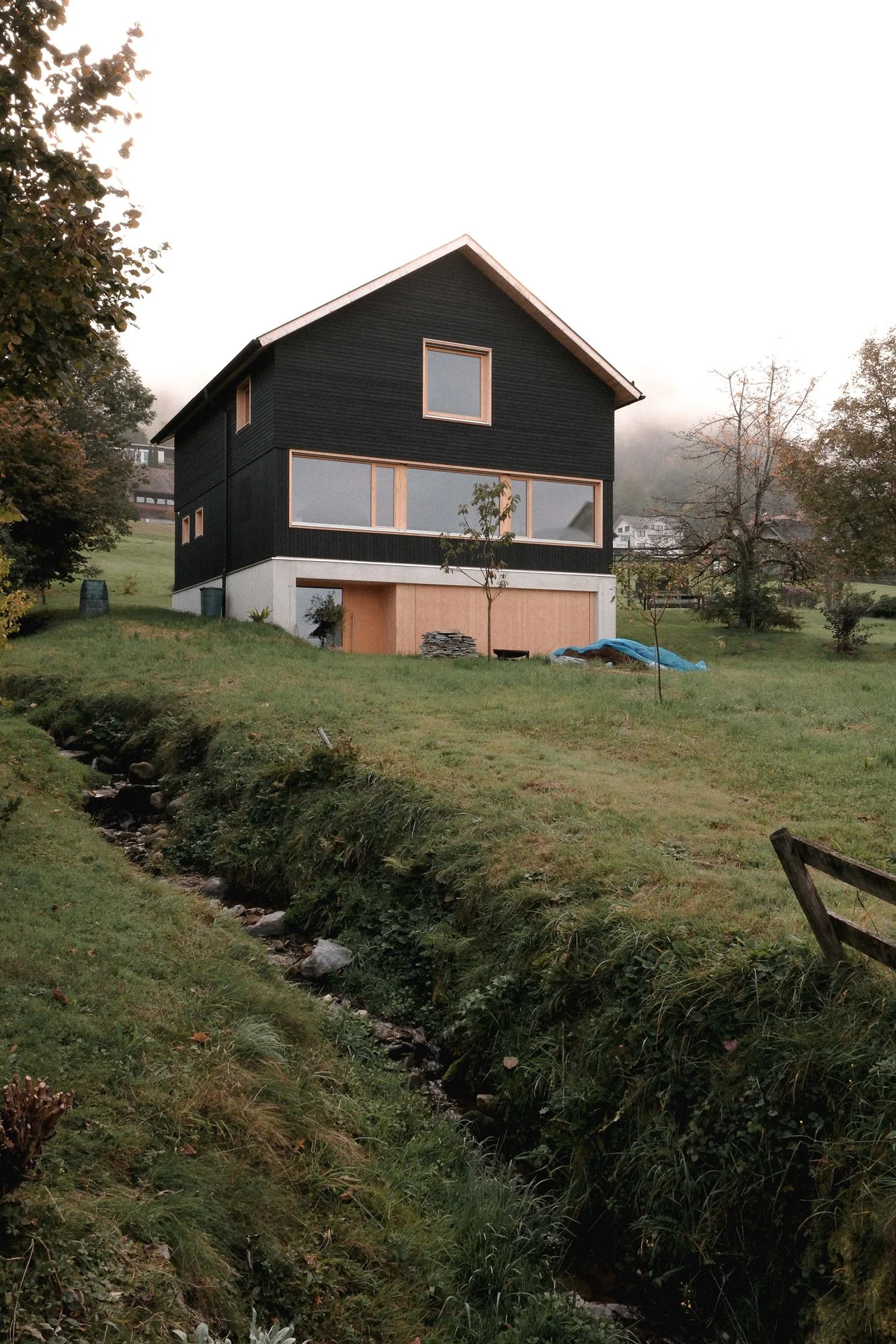 Ein modern gebautes Haus mit schwarzer Fassade, großen Fenstern und einem weißen Dach, auf einer grünen Wiese mit Bäumen und einem kleinen Bach im Vordergrund.