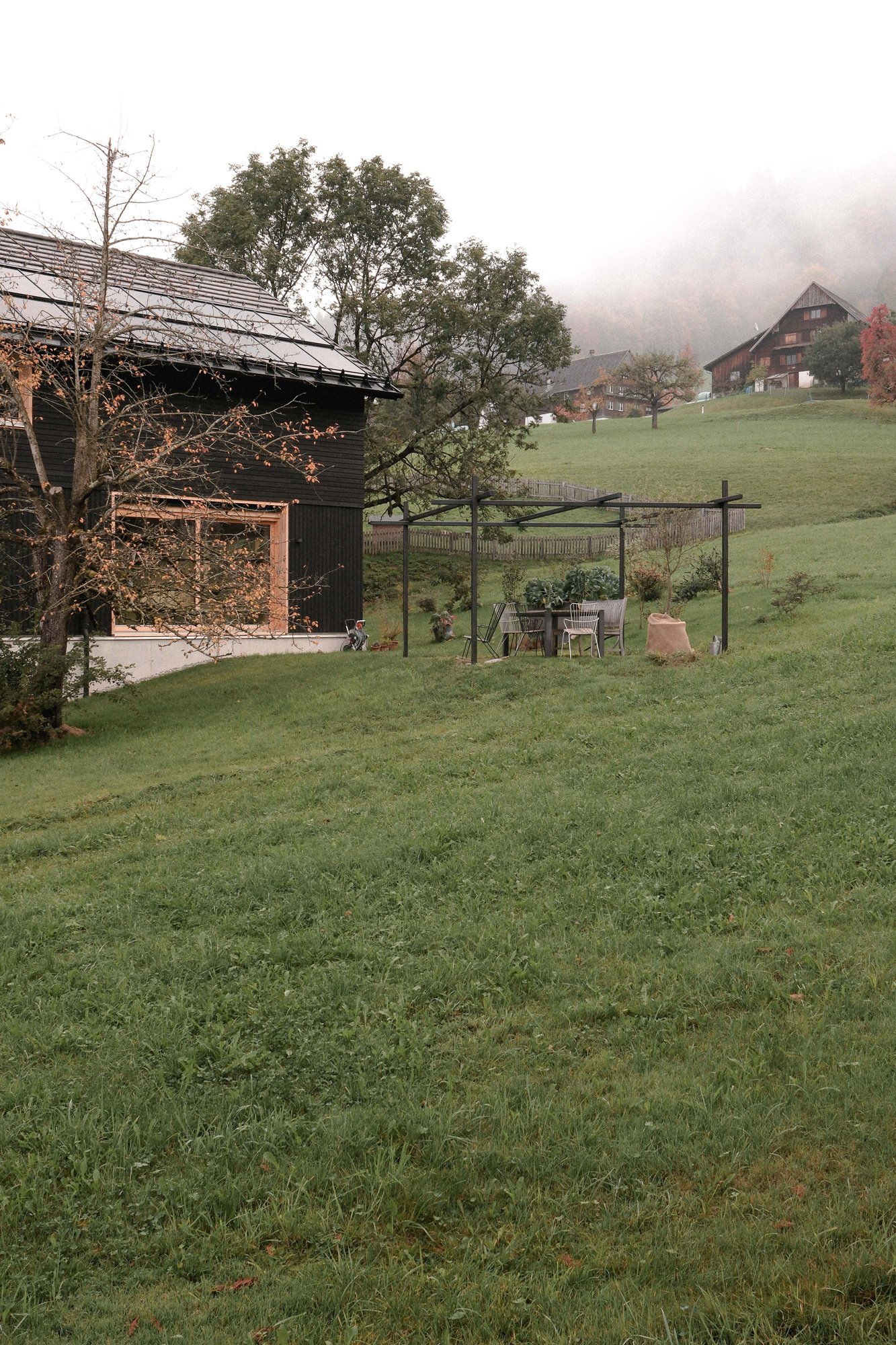 Ein Garten mit einem schwarzen Haus, einem Baum mit wenigen Blättern, einer Gartenlaube mit Sitzgelegenheiten und einem großen grünen Hügel im Hintergrund, in einer nebeligen Landschaft.