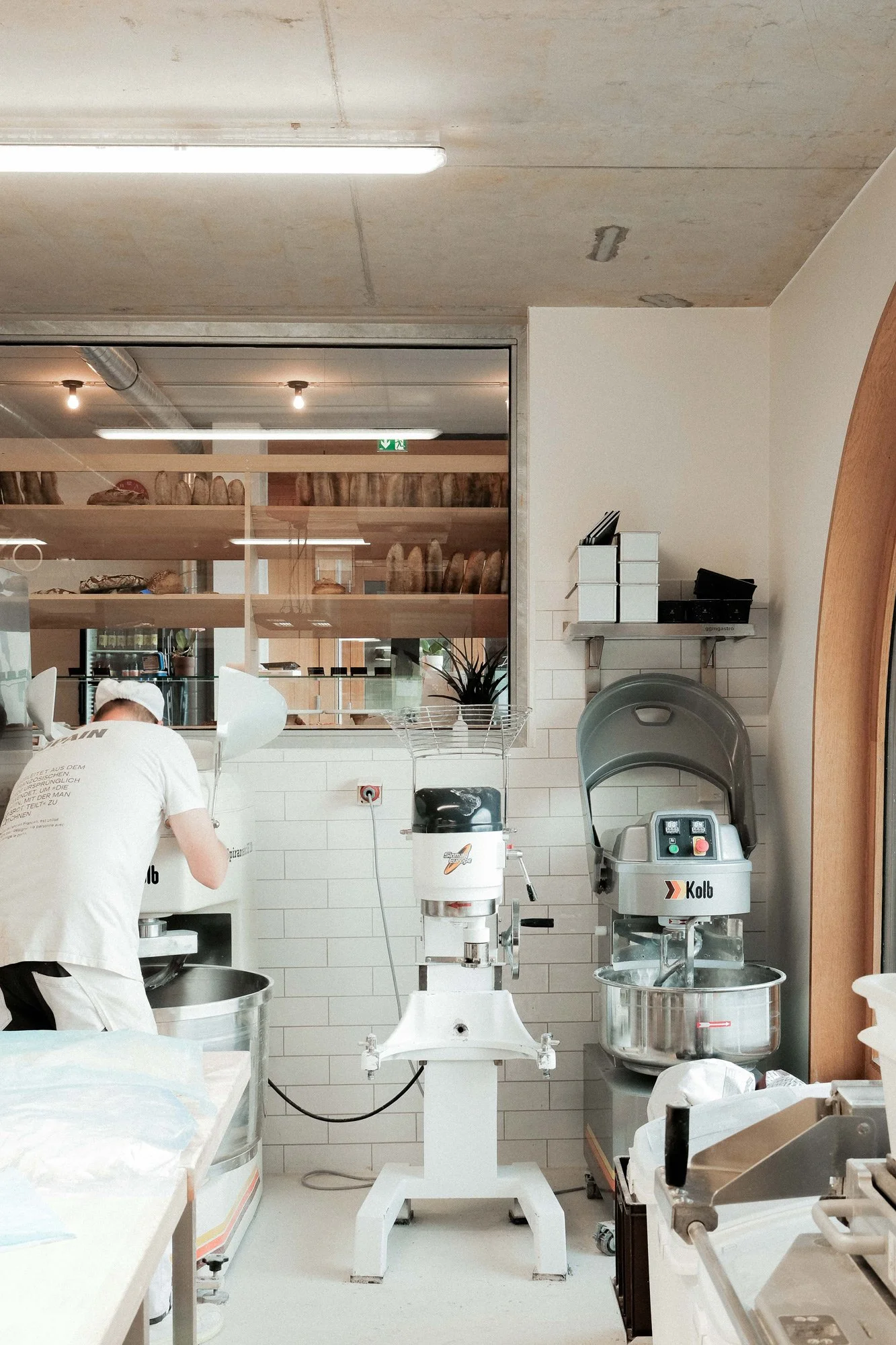 Bäckerei- oder Konditorei-Backstation mit einem Mitarbeiter, der Mehl oder Teig verarbeitet, und verschiedenen Backmaschinen, in einem modernen, sauberen Raum mit weißer Fliesenwand und Sichtfenster auf ein Lager mit Broten und Backwaren.