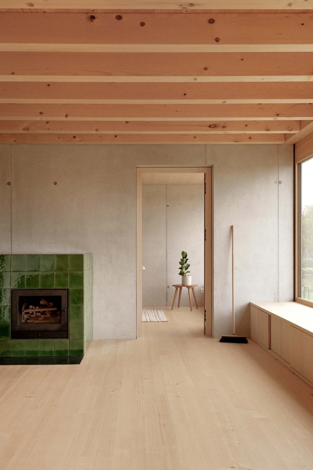 Innenraum eines minimalistischen Hauses mit Holzdecke, grün gefliertem Kamin, Holzfußboden, einem kleinen Fensterbank, einer Tür zum nächsten Raum und einem kleinen Beistelltisch mit Pflanzen.
