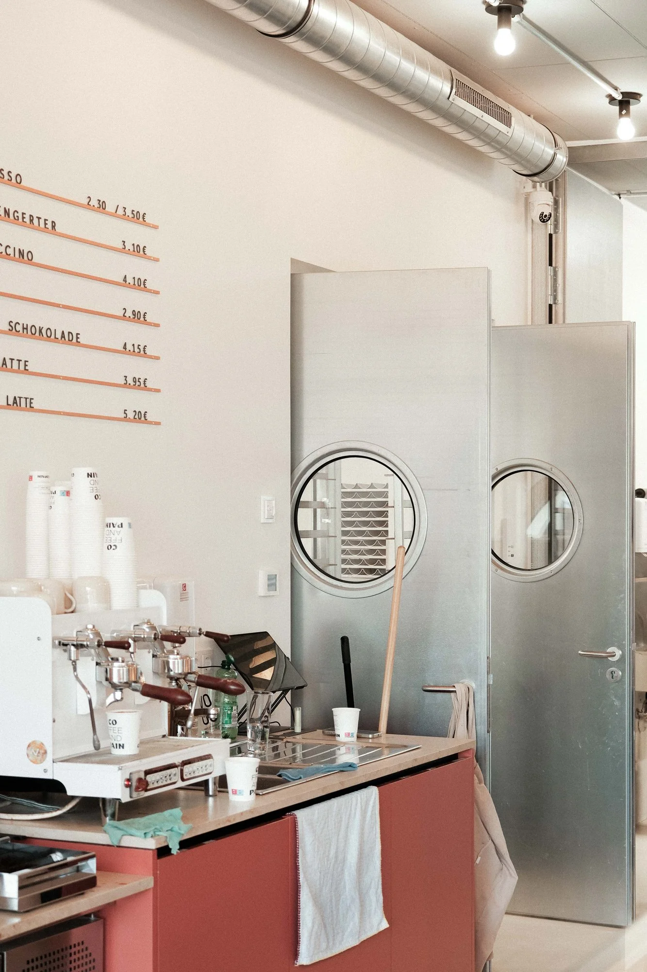 Ein Café mit einer weißen Wand, auf der die Getränkepreise in Euro auf einer schwarzen Wandtafel geschrieben sind. Es gibt eine Barista-Station mit Espressomaschine, Tassen und Getränken. In der Mitte befindet sich eine Tür mit einem runden Fenster, die wahrscheinlich zur Küche oder einem Lagerraum führt. Auf dem Tresen liegen Papiertücher und einige Tassen, während an der Wand eine Handbrause hängt.
