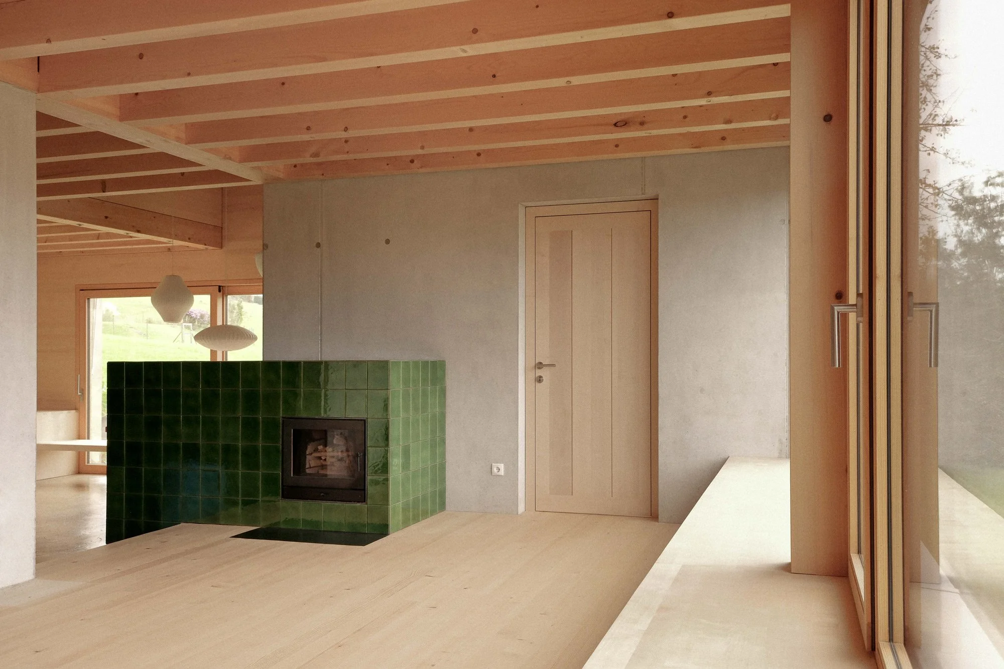 Innenraum eines modernen Hauses mit Holzdecke, grüner Kachelofen, Fenster und Tür aus hellem Holz.