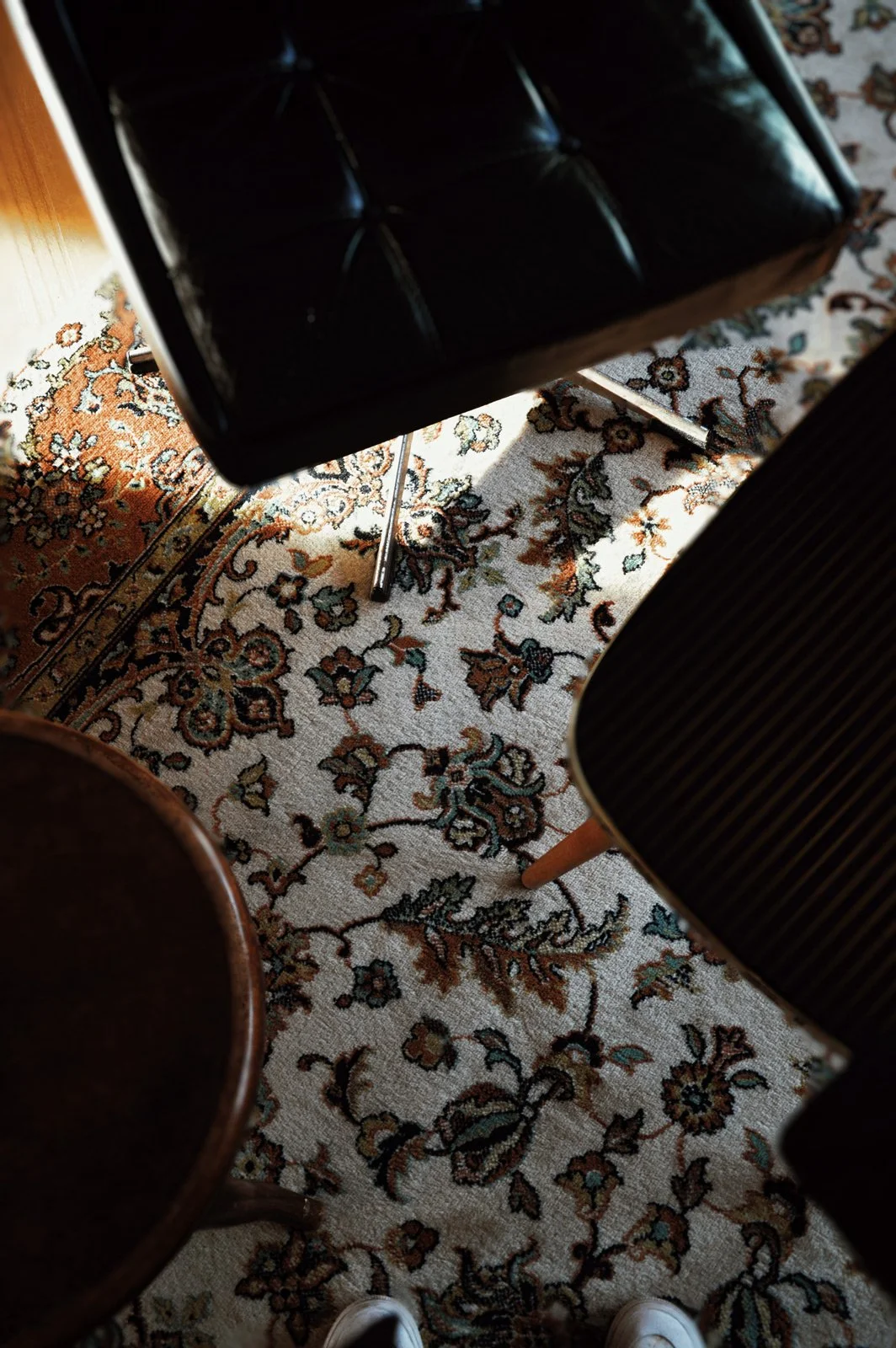 Blick auf einen bunten, orientalisch inspirierten Teppich mit drei Möbelstücken: ein schwarzer Hocker, ein Stuhl mit schwarzer Rückenlehne und ein runder Holzstuhl, sowie eine schwarze Sitzgelegenheit, alles in einem Raum mit Holz- und Teppichboden.