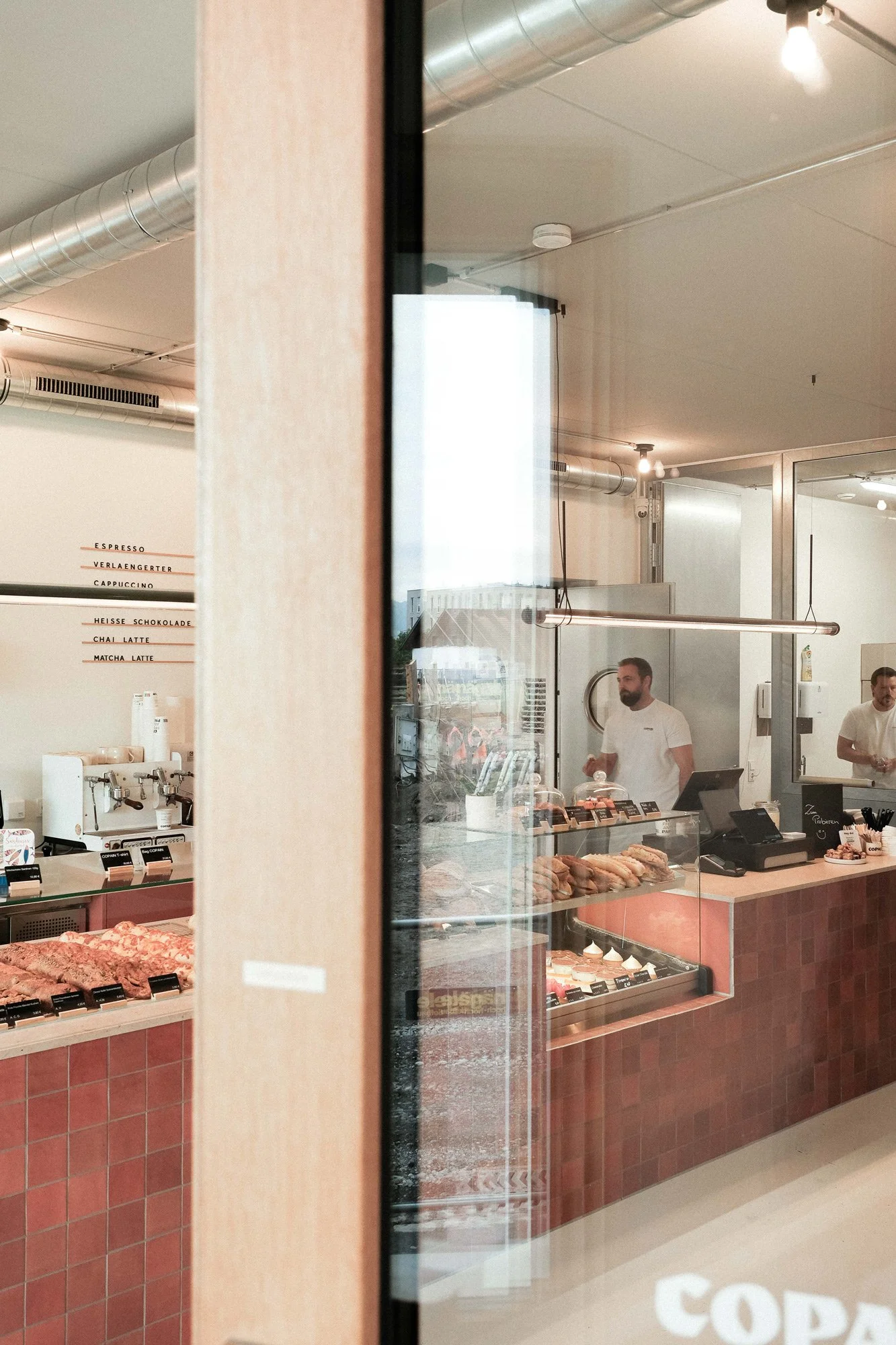 Blick in eine Fleischerei mit Theke voller Fleischwaren, zwei Mitarbeiter hinter der Theke, ein großer schwarz-weißer Kassenautomaten, im Hintergrund ein Menü mit verschiedenen Kaffeearten, durch das Fenster sind die Mitarbeiter zu sehen.