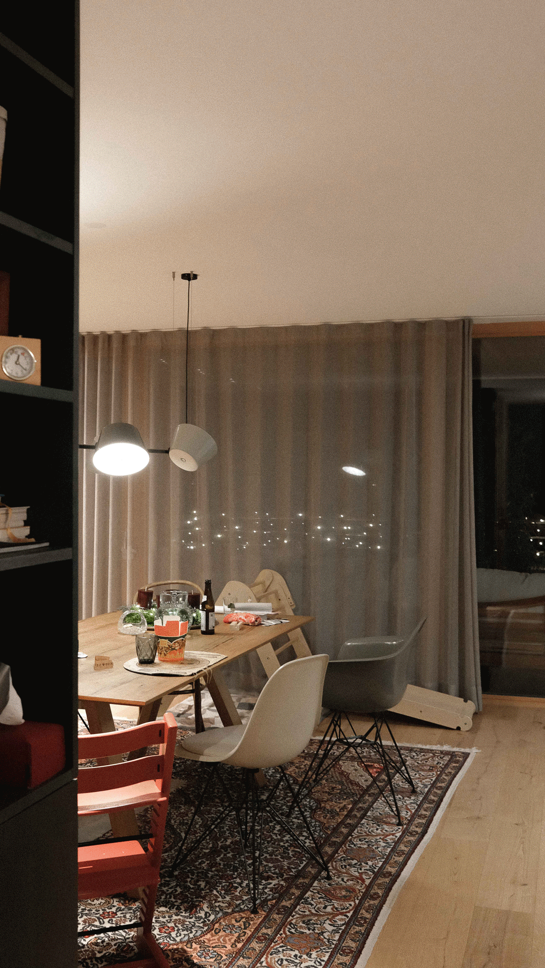 Ein Esszimmer mit einem Holztisch, verschiedenen Stühlen, einem Teppich, einer Deckenlampe, einem Fenster mit Vorhängen und Gegenständen auf dem Tisch, einschließlich Flaschen, Gläsern und Lebensmitteln.