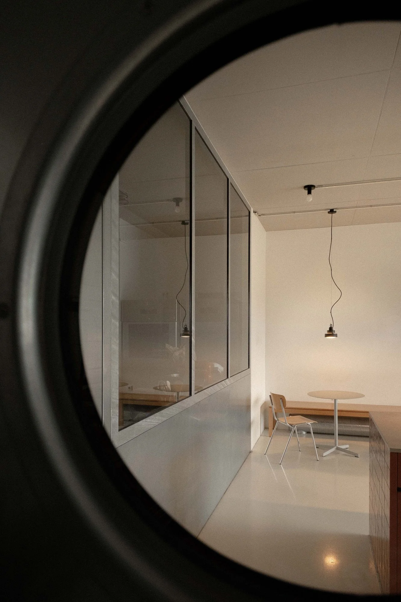 Innenaufnahme eines modernen, minimalistischen Raumes, gesehen durch ein geöffnetes Waschmaschinenfenster. Der Raum hat weiße Wände, eine runde kleine Tafel, einen Stuhl, hängende Lampen und einen Glas-Wandschrank.