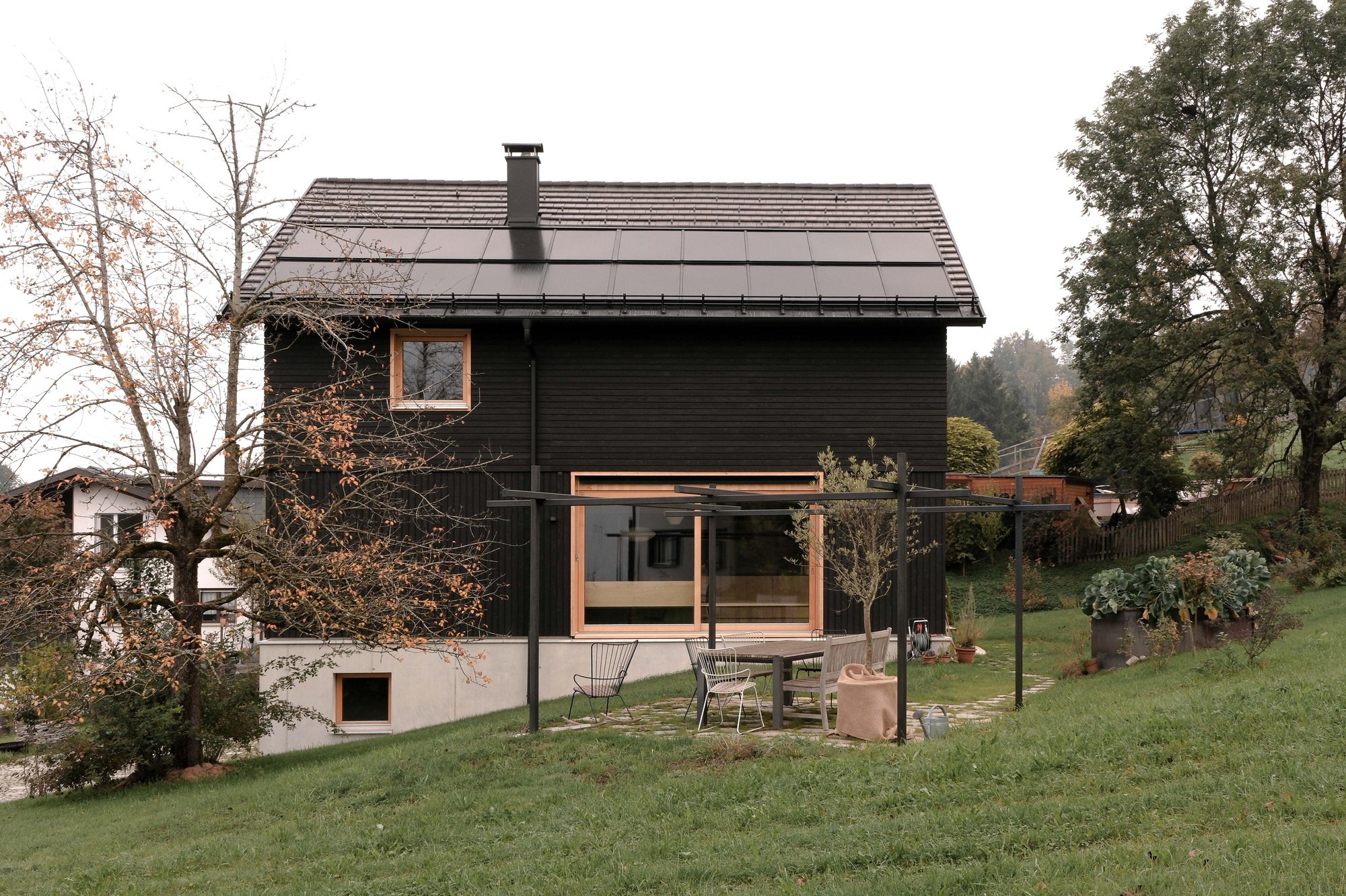 Ein dunkles Holzhaus mit Solarpanelen auf dem Dach. Im Vordergrund ein Garten mit Sitzbereich, Bänken und einem Wäscheleine. Es gibt Bäume und grüne Büsche im Garten.