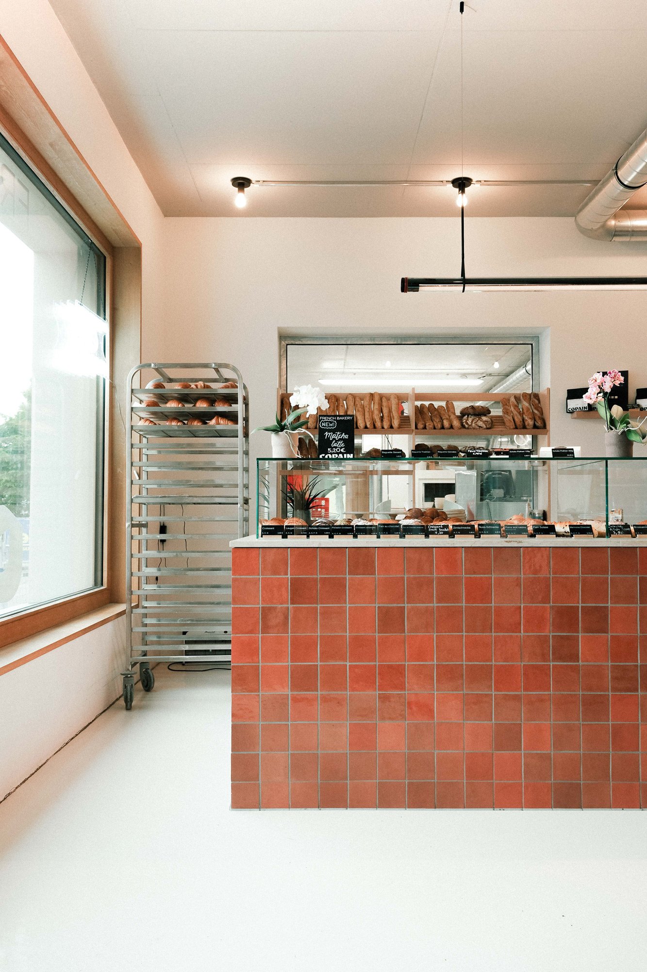 Innenraum einer Bäckerei mit einem Verkaufs-Vitrine voller Backwaren und einem Ladentresen, im Hintergrund Regale mit weiteren Backwaren, große Fenster auf der linken Seite, und ein Metallwagen für Backwaren auf der linken Seite.