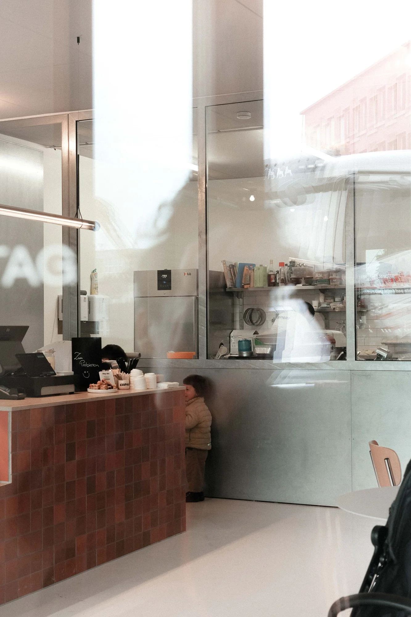 Ein kleiner Junge steht an einer Theke in einem Café, während eine Glasscheibe den Blick in die Küche zeigt.