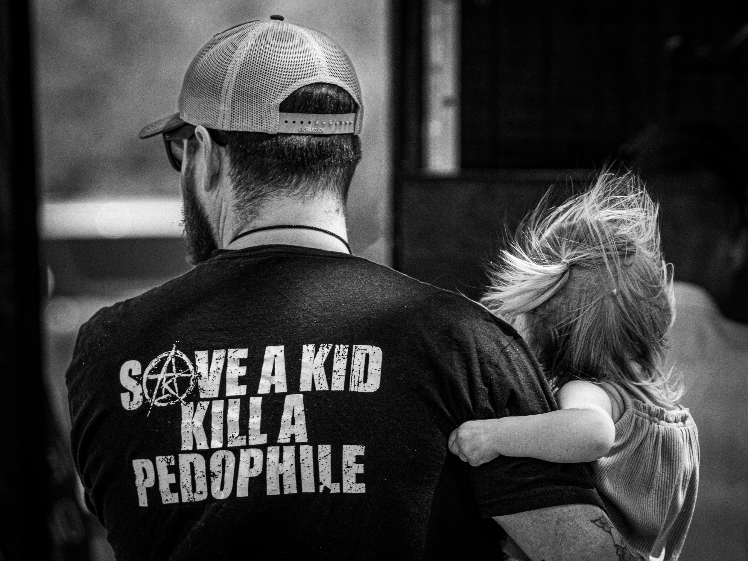 A man holding his child wearing a shirt saying, "save a kid kill a pedophile" in black and white