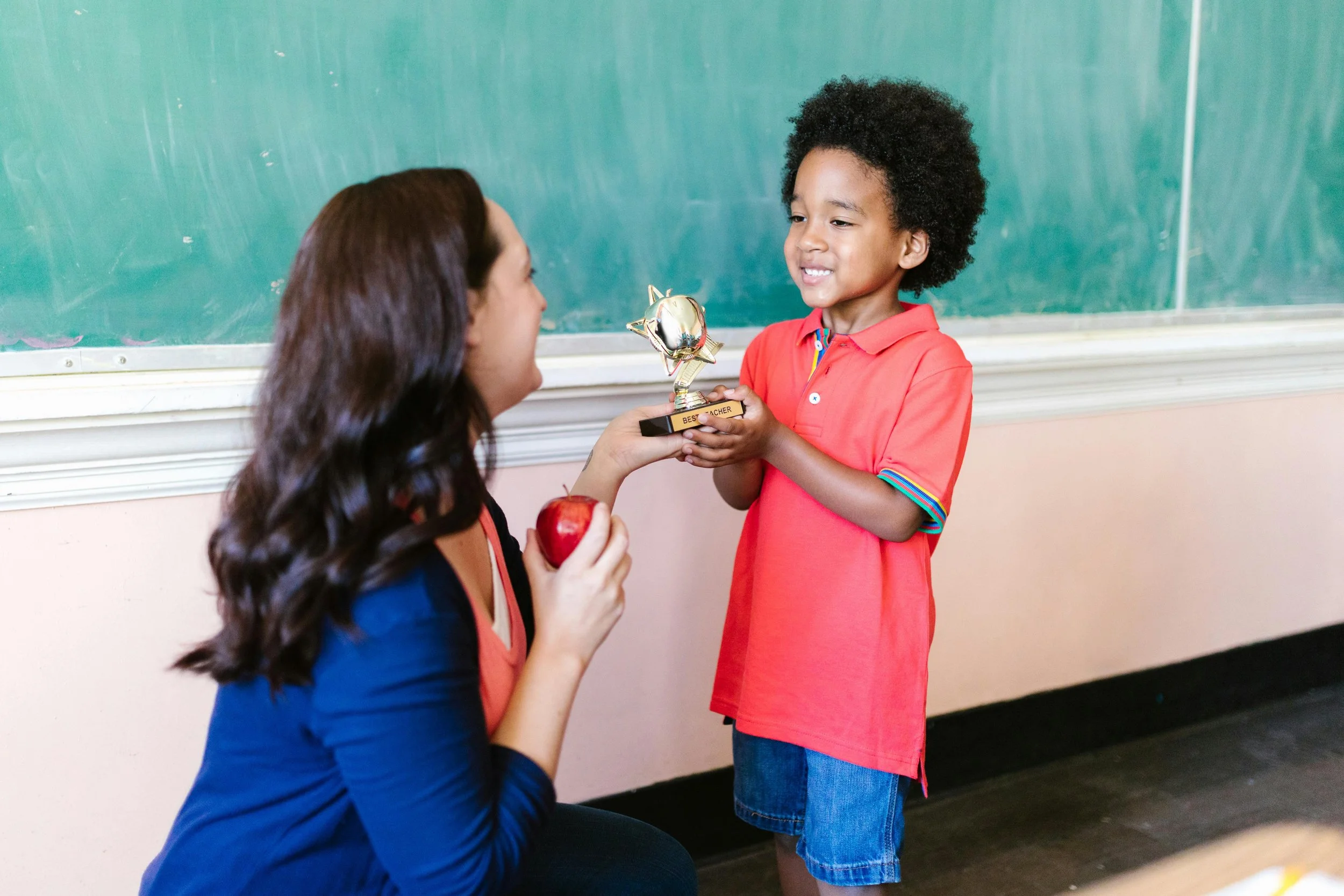 A smiling elementary school student gives his smiling classroom teacher a best teacher award and a red apple.