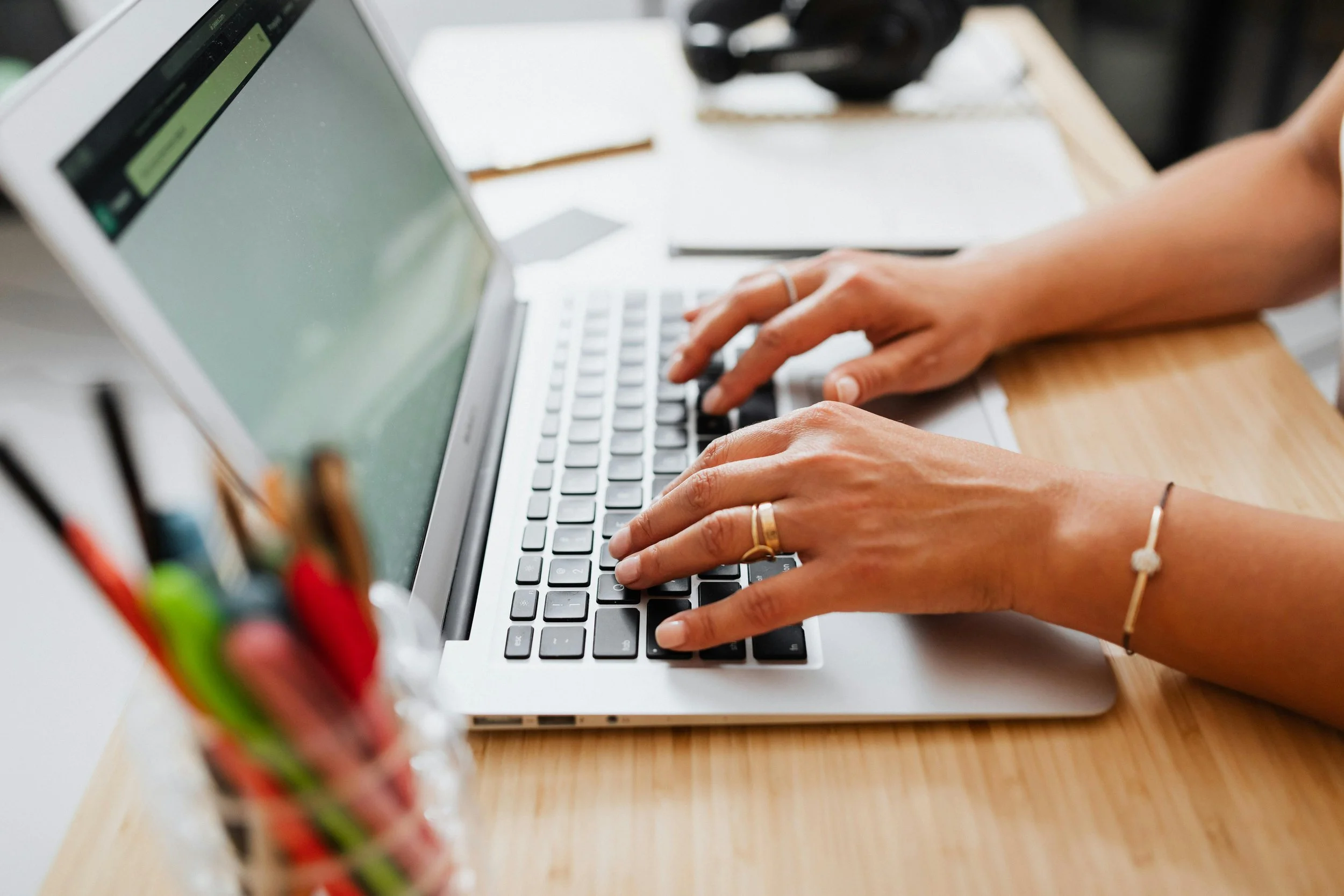 A teacher is typing on a laptop to plan lessons at a desk with a notebook, pens, highlighters, and a headset.