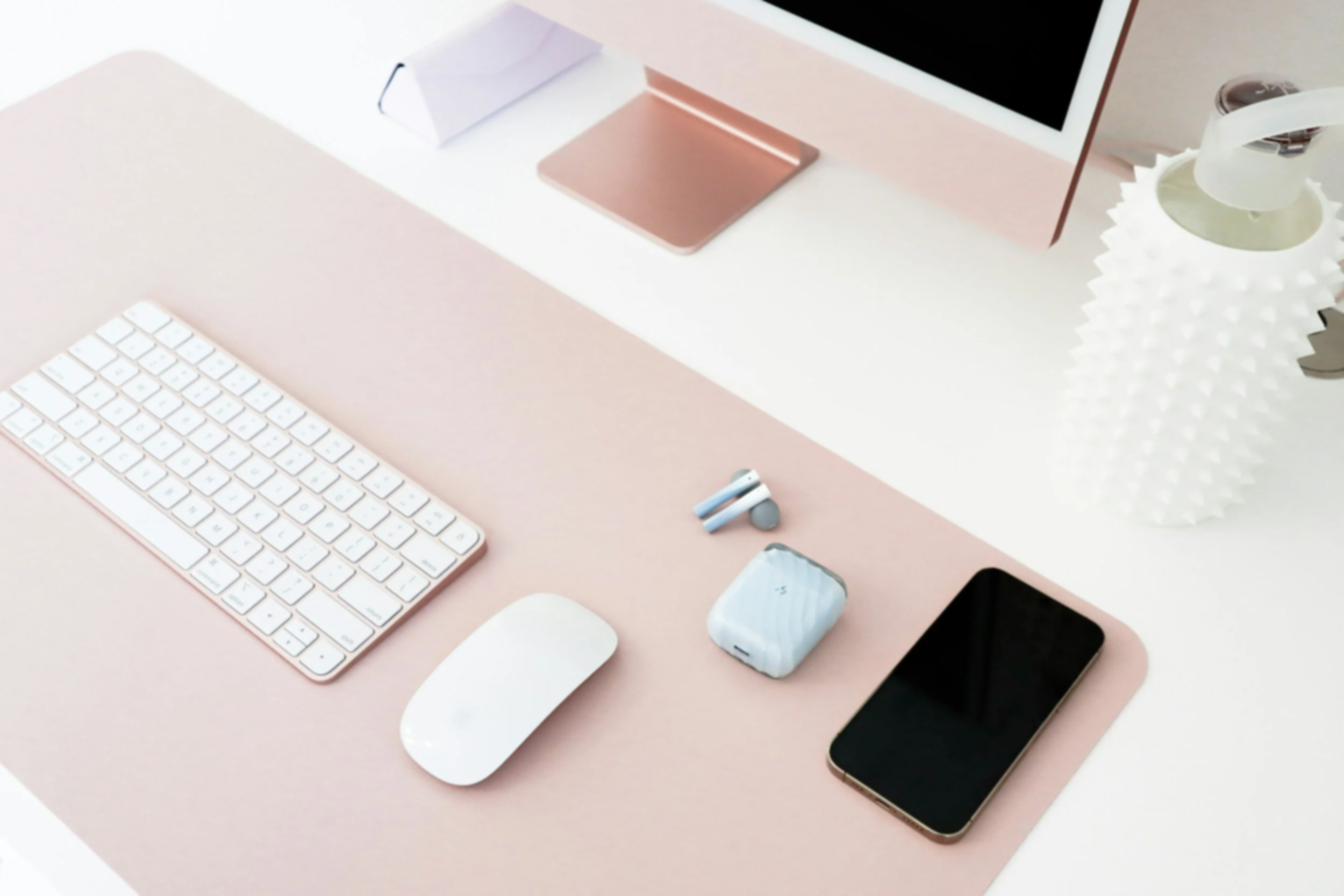 Desk with white keyboard, computer mouse, smartphone, wireless earbuds, and decorative vase on a pink and white workspace.