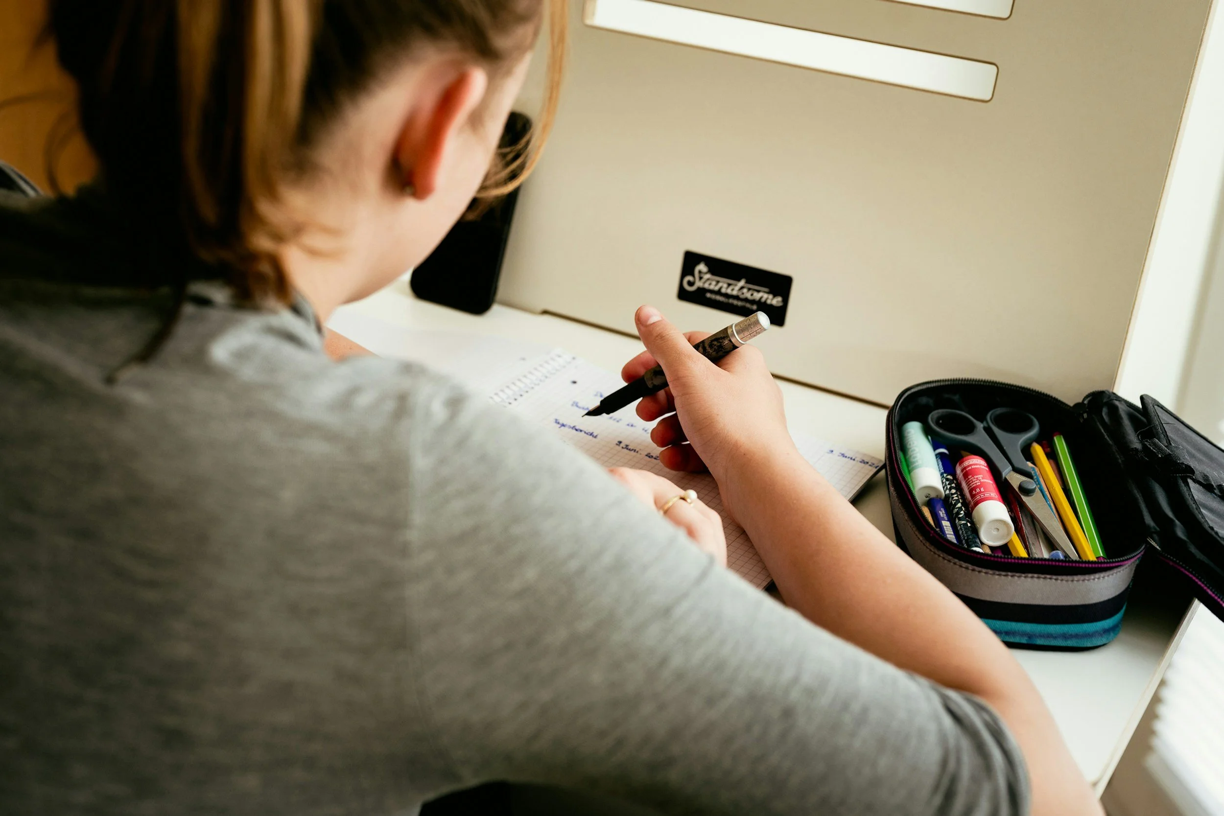 A student is sitting at a desk, working on her project. She writes in a notebook and has a supply pouch next to her.
