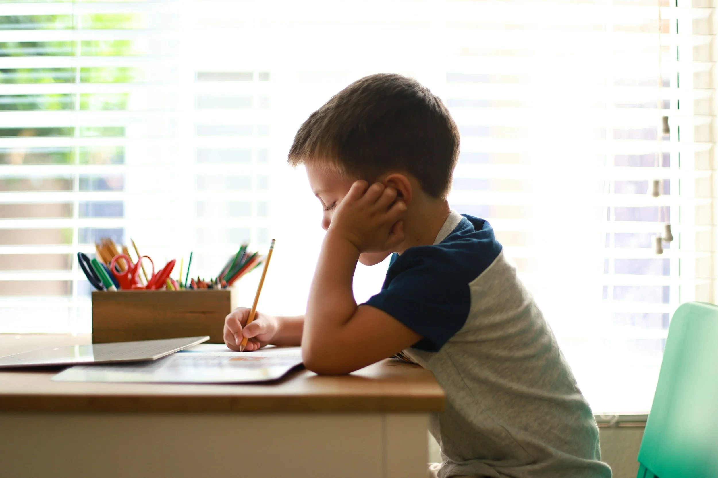 Elementary school student sitting at a desk by a window, resting his head on his hand while writing in a notebook with pencils nearby.
