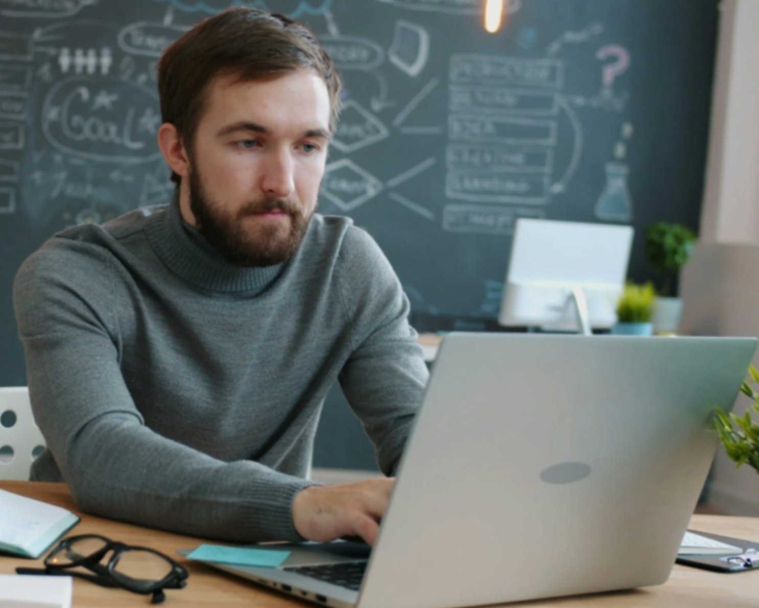 Teacher working on a laptop in a modern classroom with educational diagrams visible on a chalkboard in the background.